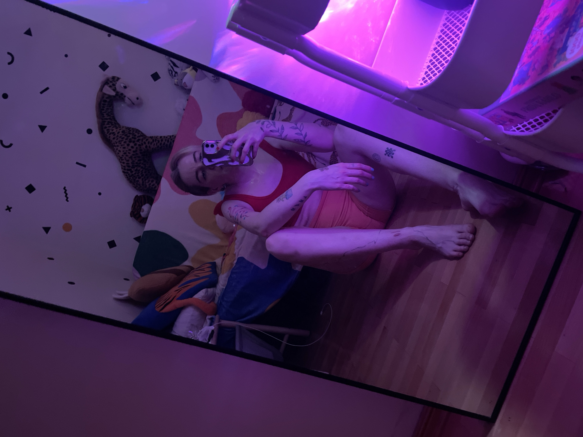 A mirror-reflected overhead shot shows a fair-skinned person with visible tattoos, sitting cross-legged on a brightly patterned blanket. The person is holding a cell phone, possibly taking a selfie. The blanket is adorned with geometric shapes, stuffed animals, and a child's chair. The surrounding environment includes a wooden floor and a pink-lit shelving unit. There is no verifiable information to confirm this photo was taken in Belgrade, Serbia.