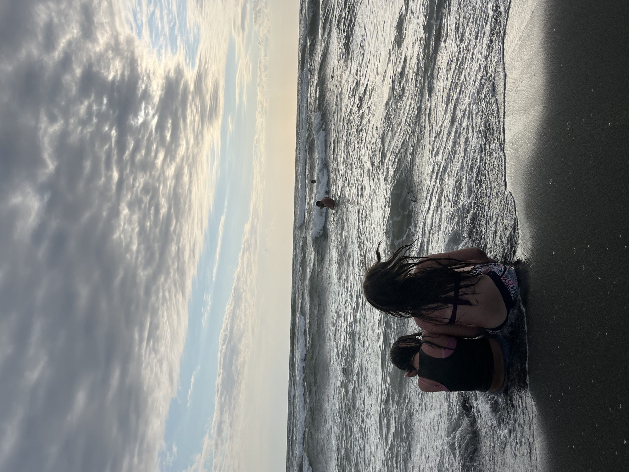 The image depicts a scene at the beach, likely in Ureki, Georgia, given the provided context. Two young individuals are sitting in shallow water near the shore, with waves gently lapping around them. The person in the foreground, facing away from the camera, is wearing a dark tank top and denim shorts. The person behind them, also facing away, has long dark hair and is wearing a patterned bikini. Their backs are to the viewer, and they appear to be engaged in a quiet moment by the sea.
In the background, the ocean stretches out, with visible waves creating a textured surface that catches the light. Further out in the water, a lone figure can be seen wading or swimming. The sky above is a dramatic expanse of clouds, with patches of sunlight breaking through, suggesting either early morning or late afternoon light. The clouds are varied in texture, with some appearing fluffy and white while others are darker and more diffused.
The overall mood of the image is serene and peaceful, with the natural elements of sky and sea dominating the scene. The soft lighting and the presence of the figures at the water's edge evoke a sense of tranquility. The wet sand and the way the water interacts with the shore are also notable details. There is no visible text in the image.
