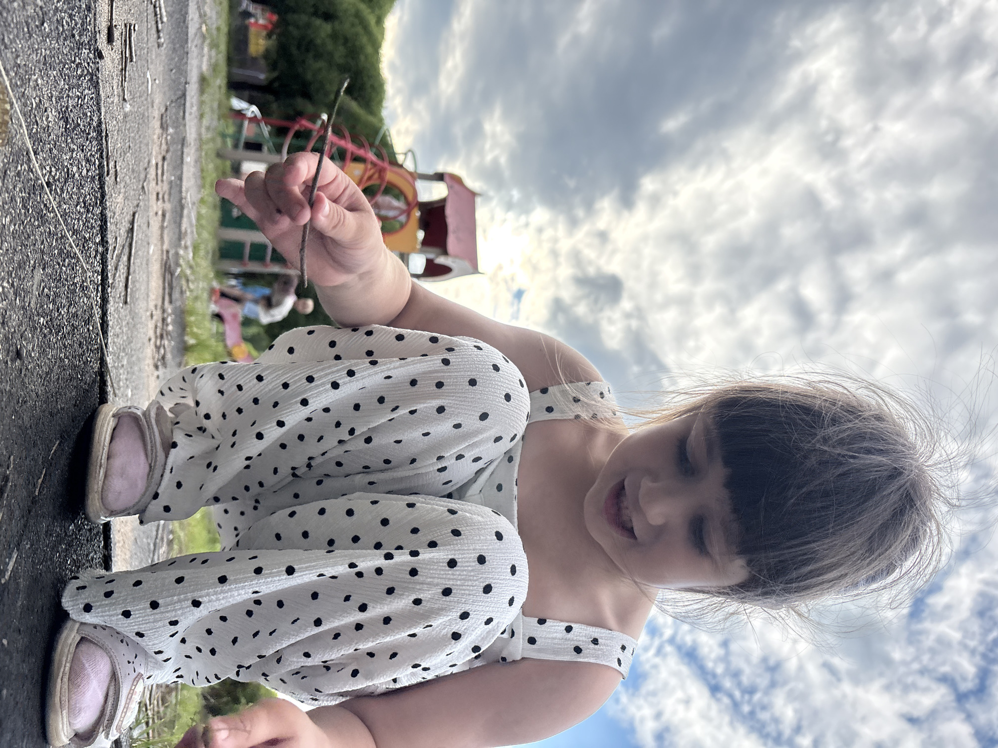 A young girl with blonde hair, wearing a white polka dot dress and pink sneakers, is crouched on a dark asphalt surface, holding a small twig in her right hand. She is looking down and smiling. In the background, a colorful playground structure with red, yellow, and pink elements is visible, along with green trees and a cloudy sky. The scene suggests it is daytime and the weather is overcast. The girl appears happy and engaged in playing outdoors, possibly in a park or playground area in Chaikovsky, Russia.