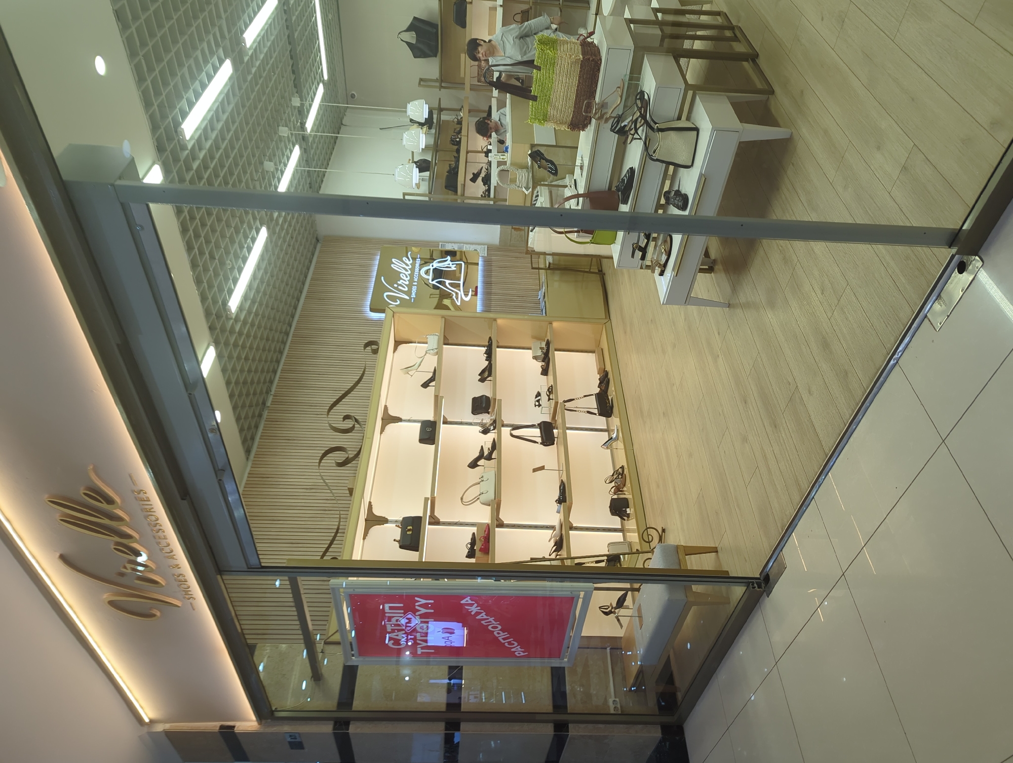 The image captures the interior of a Virelle shoe store in Bishkek City, Kyrgyzstan, likely situated in a shopping mall. The storefront is encased in glass. A red and white promotional sign written in Cyrillic, possibly advertising a sale, hangs in the window. Inside the store, there are shelves displaying handbags and shoes. Two individuals are visible; one is near the front of the store, possibly a staff member, and another is further back, presumably a customer. The interior design features a light color palette, with light wood floors and shelves. The ceiling is a white grid pattern with fluorescent lighting. The store’s name, "Virelle," is displayed in a stylized gold font on the upper part of the storefront. The floor outside the store is tiled in a light color.