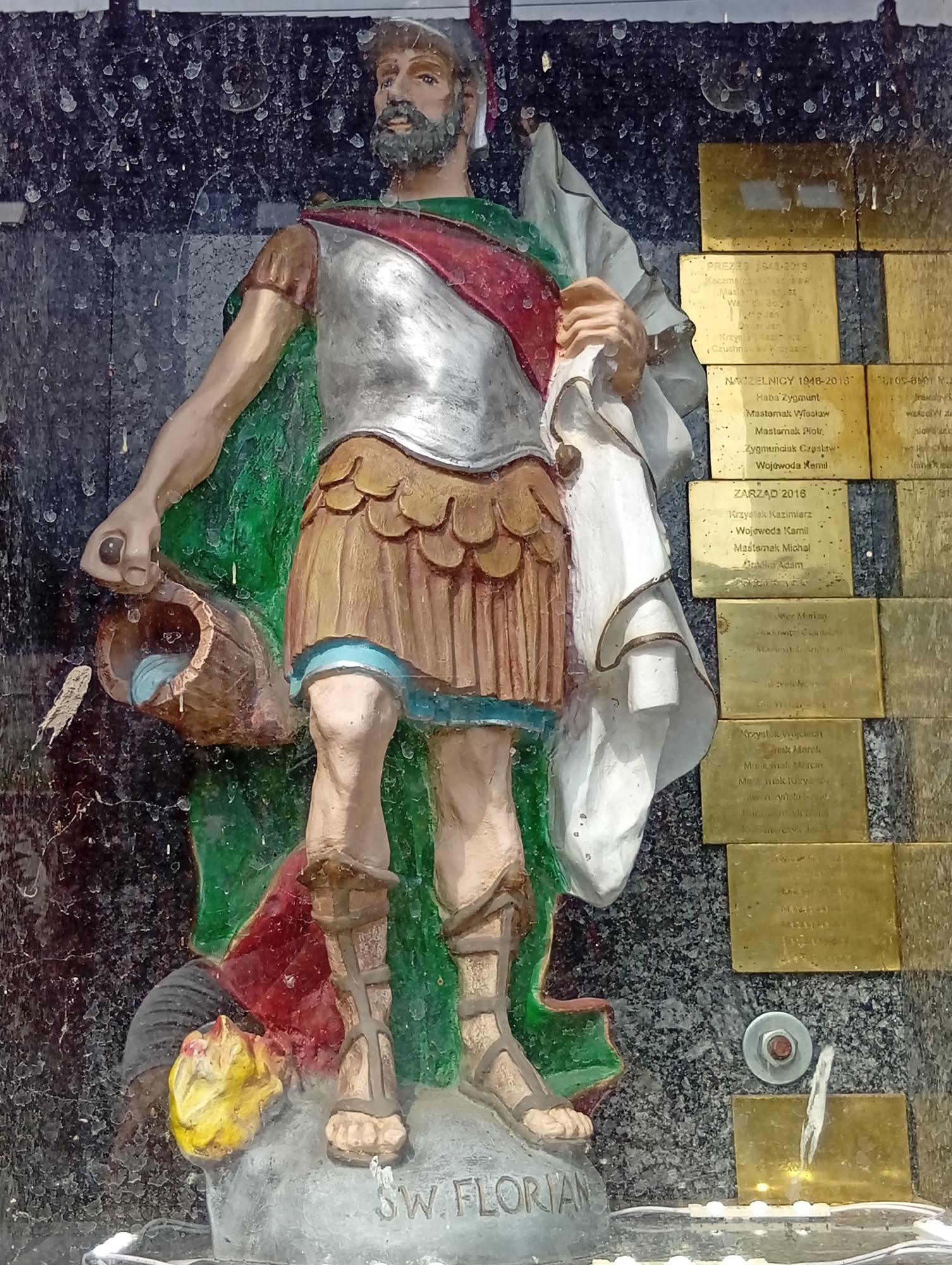 The image shows a statue of Saint Florian, a Roman soldier and martyr, holding a bucket of water. The statue is located in Brzezia Łąka, Poland, on a wall with several brass plaques. The plaques appear to be memorial or dedication plates, with names and dates inscribed on them. The statue is colorful, with Saint Florian wearing a silver cuirass, a brown skirt, and a red and white sash. He is also wearing sandals. The background is a dark, speckled wall, possibly granite, and there are some water droplets or stains on the surface. The statue is placed on a stone base with the inscription "ŚW. FLORIAN" visible. The overall scene suggests a religious or historical monument.