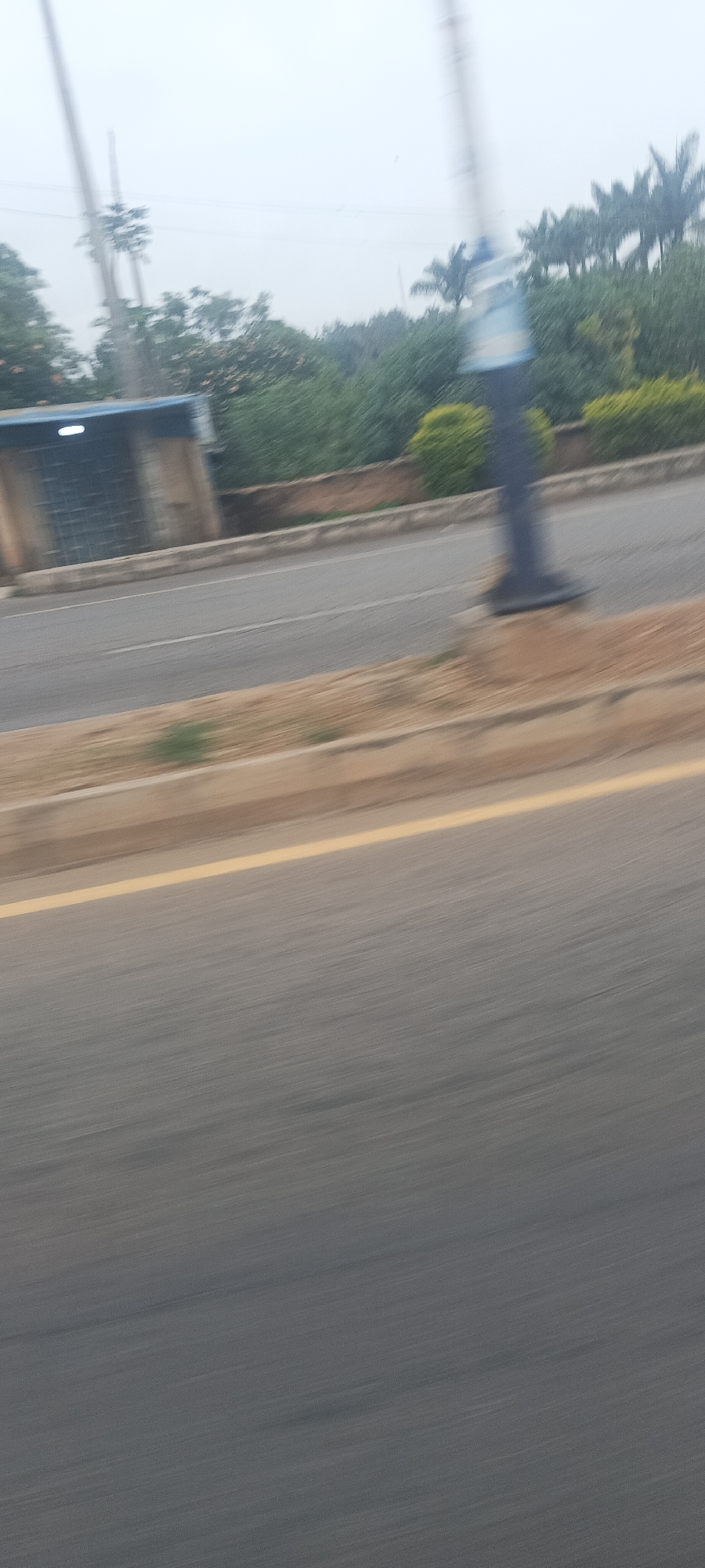 The image captures a blurred street scene, likely taken from a moving vehicle, under an overcast or hazy sky. The foreground shows an asphalt road, partially marked by a solid yellow line delineating a lane or shoulder. Adjacent to the road is a strip of reddish-brown earth or sand, followed by a low concrete barrier or curb.
Beyond the barrier, lush green vegetation forms a dense backdrop, comprising various trees and bushes, including several tall palm trees. On the left side of the frame, a small, rectangular building or structure with a dark facade and a blue roof or awning is visible, featuring a bright light source underneath its roof. This could be a bus stop shelter or a small commercial kiosk. Towards the right, a tall, dark, cylindrical pole, possibly a street light or utility pole, stands vertically, with a lighter-colored, possibly blue, component near its top.
The significant motion blur throughout the image suggests movement during capture. No people or specific activities are clearly discernible, and there is no visible text. The scene depicts a typical road environment in what appears to be an urban or suburban setting, consistent with areas like Jos, Nigeria, given the natural foliage.