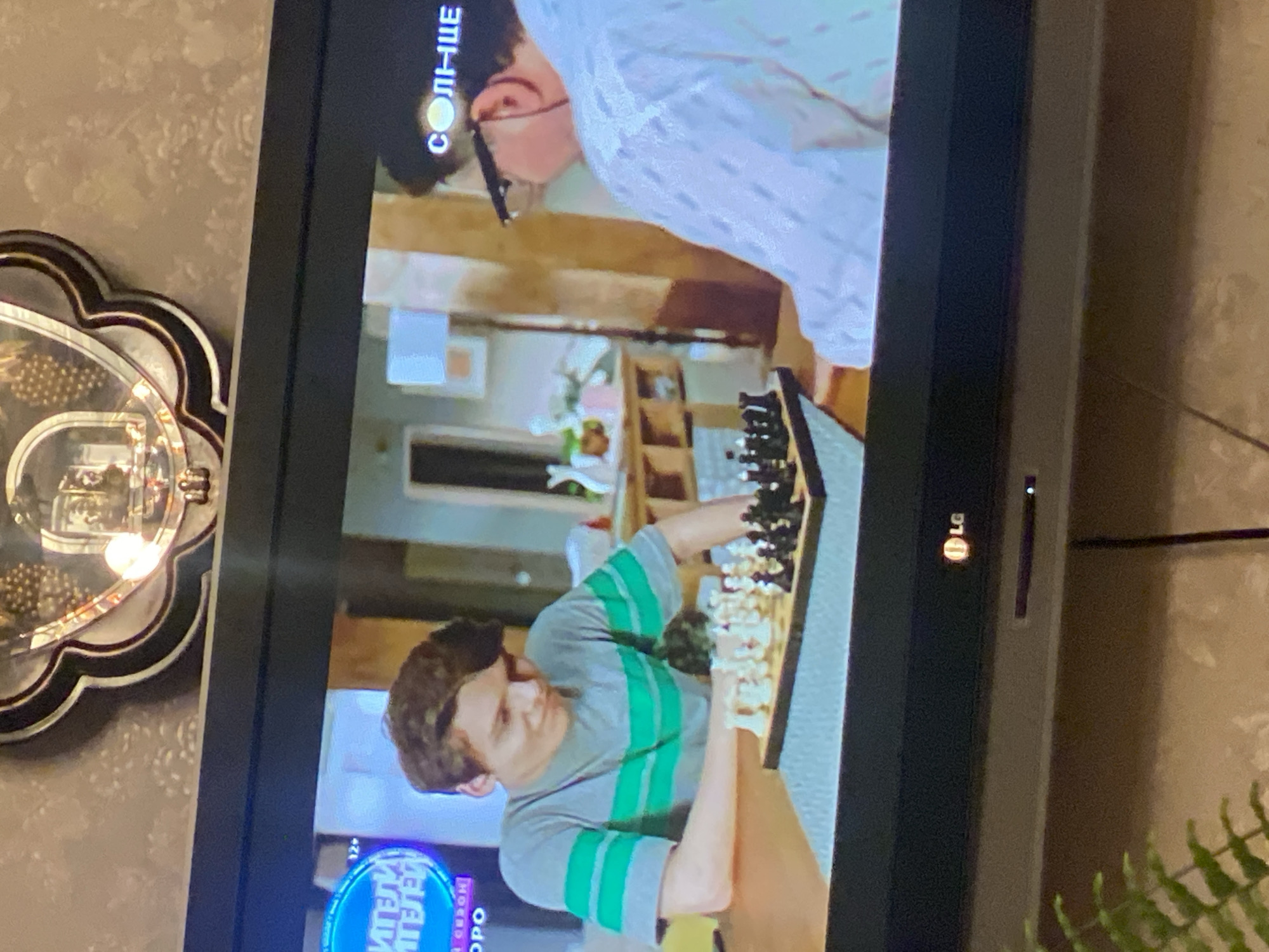 The image shows a television screen displaying a scene with two people playing chess. The person on the left is a young male with brown hair, wearing a grey t-shirt with horizontal green stripes. He is looking down at the chessboard with a slight smile. The person on the right is partially visible, wearing glasses and a white, textured shirt. They are also looking towards the chessboard.
The chessboard is set up on a table in front of the younger male. The chess pieces are arranged for a game, with white pieces on one side and black on the other. The background of the scene on the television shows a room with furniture and decor, including a lamp and a potted plant. There are also on-screen graphics, including a blue oval with text that is partially obscured, and the word "СОЛНЦЕ" (which translates to "SUN") with a circular graphic above it.
The television itself is dark around the edges, indicating it is a modern flat-screen model. A logo for "LG" is visible in the bottom center of the frame, along with a small blue indicator light. The television is positioned in a room with patterned wallpaper on the wall behind it and a decorative mirror frame above the screen. A potted fern is visible in the lower left corner of the image, in front of the television. The overall impression is that of a casual setting, possibly a living room. There is no information in the image that can be used to determine the location in the city of Novaya, Russia.