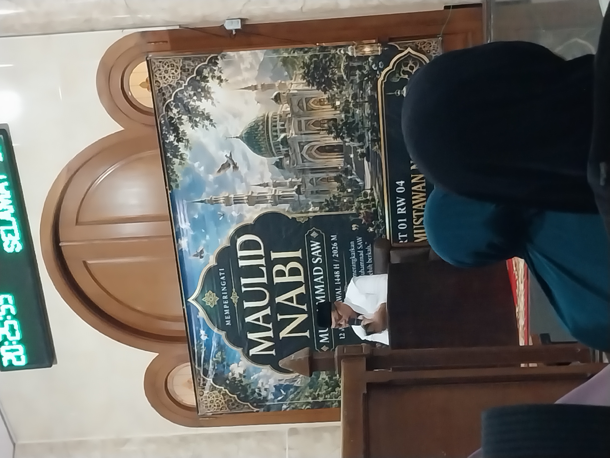 A man in a white shirt and black cap, wearing glasses, is speaking into a microphone. He is standing behind a dark brown podium with a wooden frame. The man is in front of a large banner that reads "MEMPERINGATI MAULID NABI MUHAMMAD SAW". The banner also features an illustration of a mosque and Islamic decorative patterns. To the left of the banner, there is a digital clock displaying "20:25:55" and "SELAMAT". The scene appears to be indoors, possibly in a mosque or community hall in Nglegok, Indonesia. In the foreground, there are the backs of several people wearing headscarves. The overall lighting is dim, with the banner and the digital clock being the brightest elements.