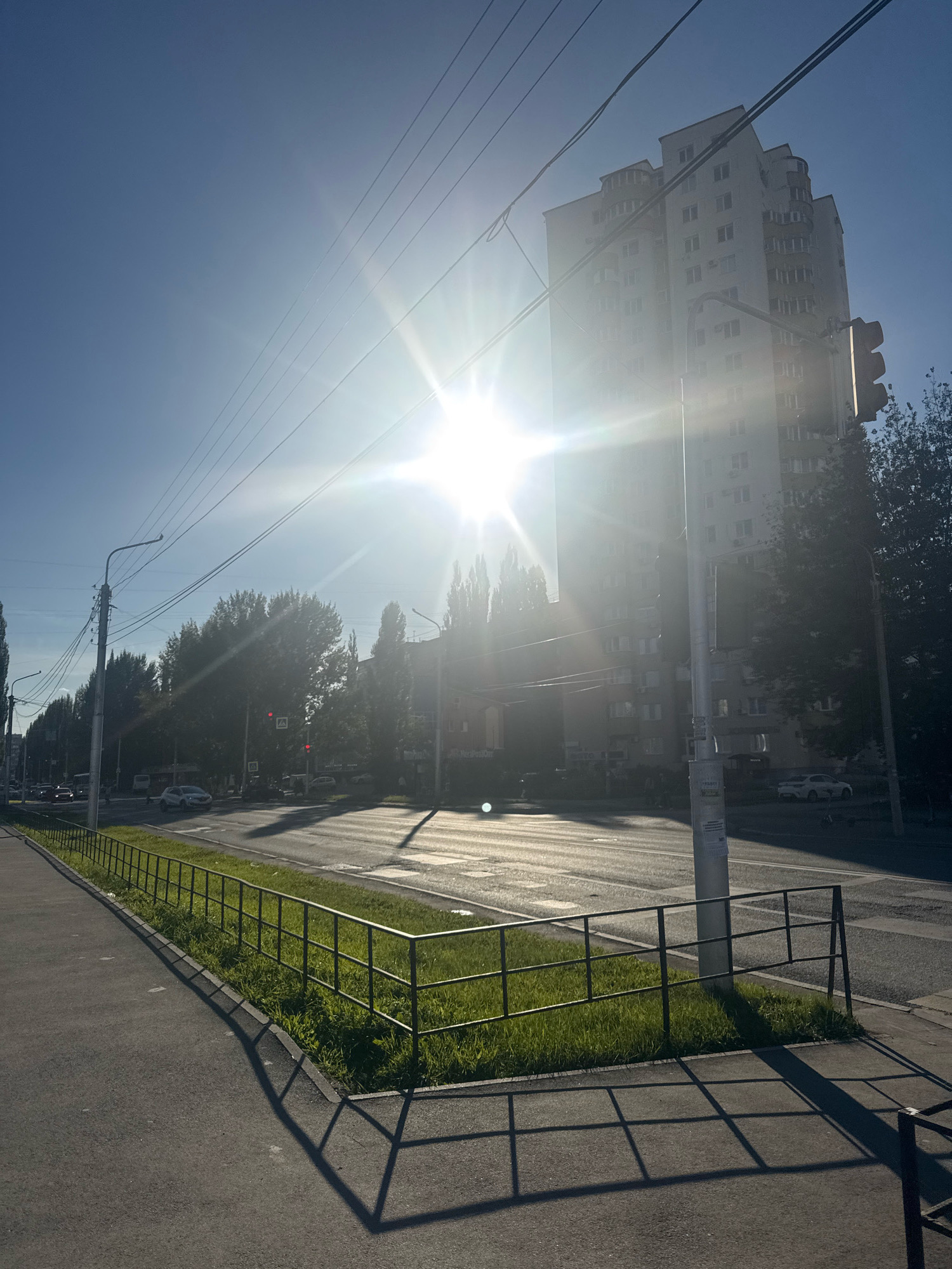 The image captures a bright, sunny day on a street in Ufa, Russia. The sun is high in the sky, creating lens flare and strong shadows. In the foreground, a paved sidewalk runs alongside a grassy median bordered by a black metal fence. The fence casts a geometric shadow onto the sidewalk. Across the street, a multi-story apartment building with numerous windows dominates the background. A traffic light, trees, and other urban infrastructure, including power lines and streetlights, are visible. Cars are present on the road, some moving and others parked. The overall impression is of a typical urban street scene bathed in strong sunlight.
