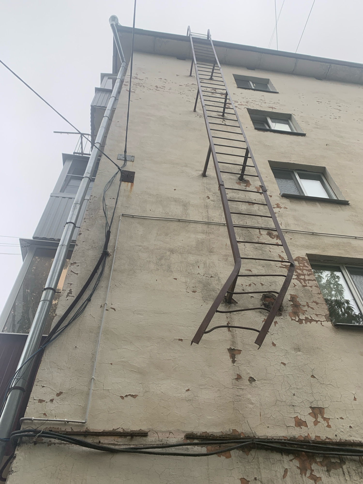 An exterior, low-angle, full shot captures a weathered, multi-story building in Arkhangelsk, Russia, under an overcast sky. A tall, rusty, metal fire escape ladder extends vertically along the beige, peeling facade, with its lower section bent outwards. Several windows are visible on the building's surface, some reflecting the cloudy sky and surrounding trees. A vertical metal pipe, likely a downspout, runs along the left side of the building, adjacent to a balcony and other pipes and electrical wires. The overall condition of the building's exterior suggests age and disrepair, with significant cracking and flaking of the paint. The environment is characterized by a muted color palette and a sense of urban decay. There are no people visible in the image.