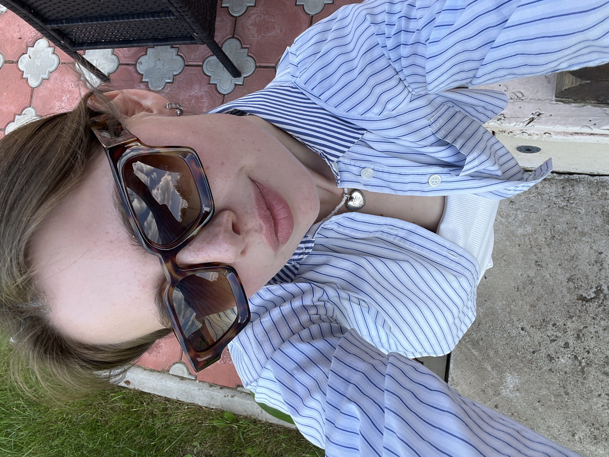 A young woman is depicted in a high-angle selfie photograph taken outdoors in Chaikovsky, Russia. She has light brown hair and wears large, square-framed tortoiseshell sunglasses that reflect a cloudy sky and distant trees. Her nose and cheeks display visible freckles, and her lips are colored with light pink lipstick, curving into a subtle smile. She wears a light blue and white striped long-sleeved collared shirt, unbuttoned to reveal a white tank top underneath. A white beaded necklace with a silver heart-shaped locket rests on her chest. A small silver hoop earring is visible in her right ear. The ground directly beneath her is rough concrete pavement. To her left, green grass meets a concrete curb. To her right, a distinctive pink-patterned tile pathway with a repeating quatrefoil design is visible, alongside a partial view of a dark, possibly woven, outdoor chair.