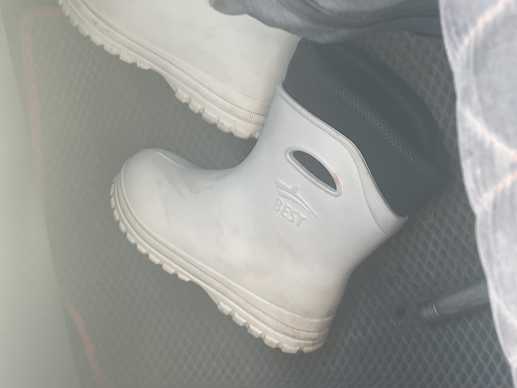 The image captures two white, short boots, likely made of rubber or PVC, stored within what appears to be a vehicle's cargo space. The boot in the foreground is prominently displayed, showing its robust, ridged sole and a black, mesh-textured lining around the ankle opening. The word "BEST" is embossed on the outer side of this boot. A second identical boot is partially visible in the upper left corner, showcasing its matching sole.
The boots are resting on a dark-colored, textured surface, possibly a rubber mat with a diamond or hexagonal pattern, typical of car trunks or floor liners. To the right, a crumpled piece of light-colored fabric, possibly a blanket or tarp, with faint reddish-grey plaid lines, partially obscures the view. A slender metallic rod or pipe is also visible near the bottom right corner of the frame.
The lighting is soft and diffused, suggesting an indoor setting or overcast conditions, and there is no indication of the time of day or specific weather. The scene depicts the boots in a state of storage, with no active event occurring. The image focuses on the objects and their immediate surroundings, conveying a practical, utilitarian context, possibly within a vehicle in Russkie Kirmeni, Russia.