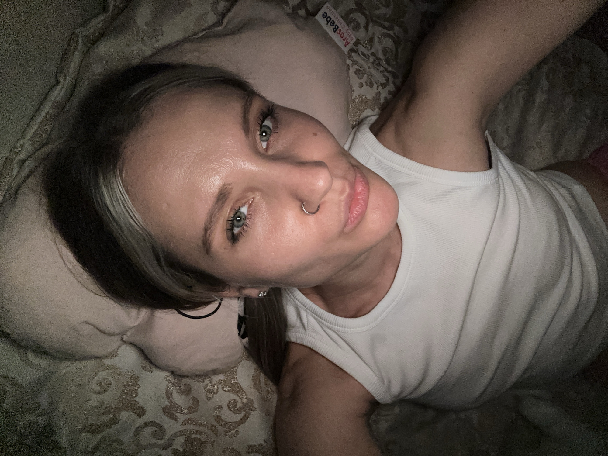 A light-skinned woman with gray and brown hair, green eyes, and a nose ring is lying on a pillow. She is wearing a white tank top. The pillow has a label that reads "ArosBebe Baby Collection." The background shows a patterned blanket or bedding. There is no information to place the image in Esenyurt, Turkey.
