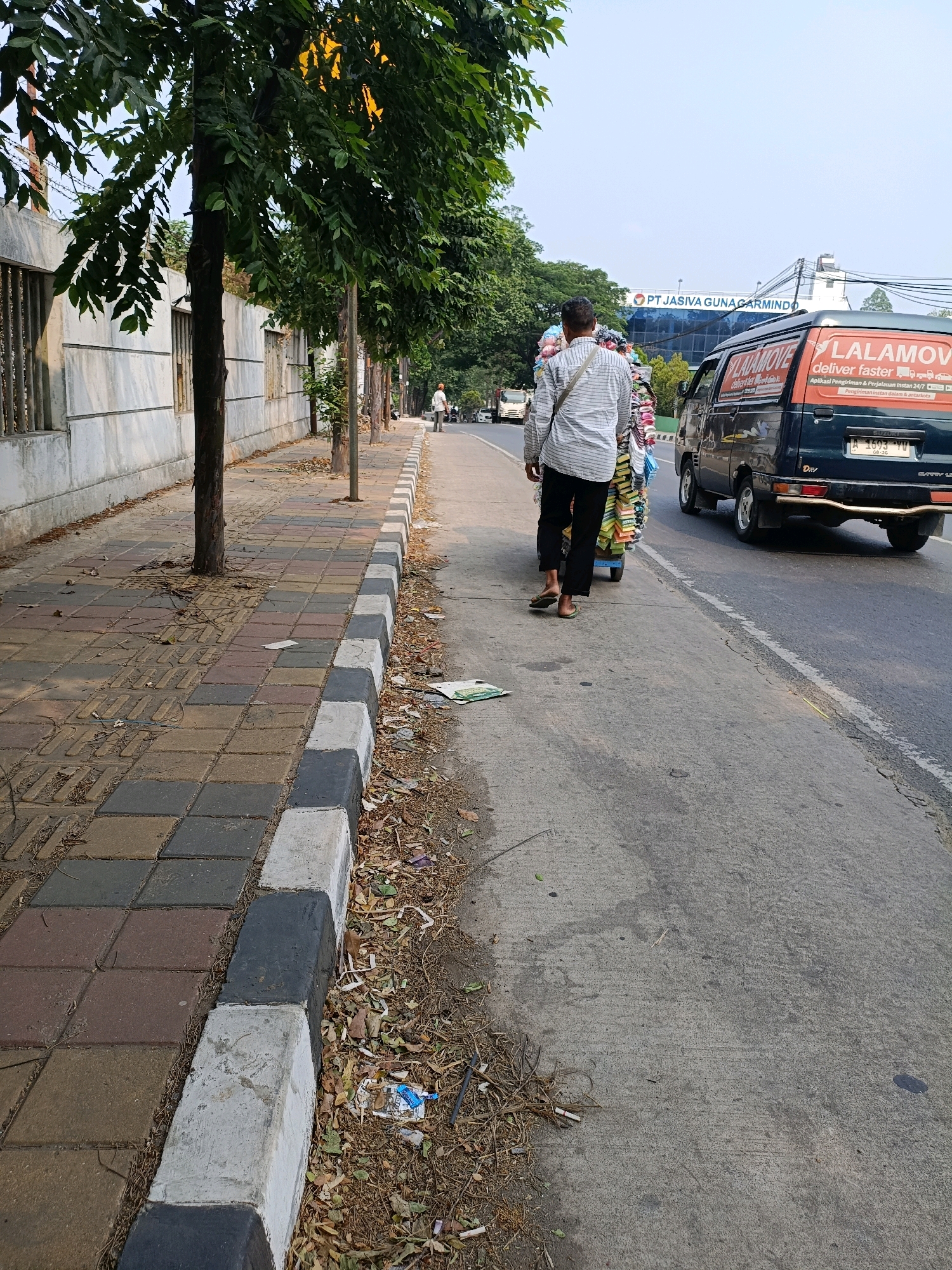 A person walks along a sidewalk in Tangerang, Indonesia, pushing a cart laden with goods. The person is wearing a striped shirt and dark pants, with their back to the camera. The sidewalk is paved with interlocking bricks and bordered by a curb painted in black and white stripes. Trees line the sidewalk, casting shadows on the ground. Beyond the sidewalk is a road with a dark blue van on it, which has "LALAMOVE" written on its side. In the background, a building with the text "PT JASIVA GUNAGARMIND" is visible. The scene appears to be in an urban or semi-urban setting.
