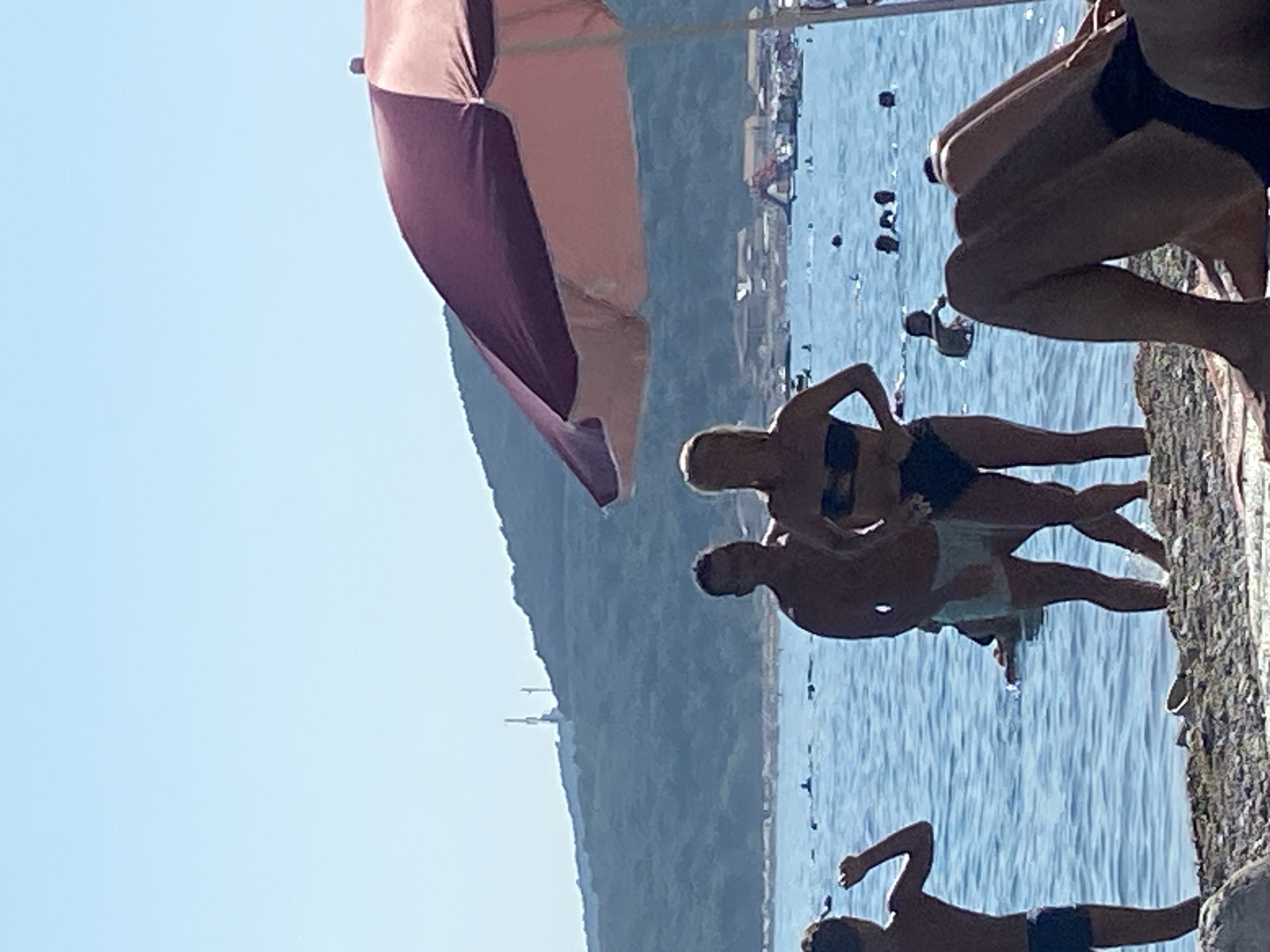 The image depicts a sunny day at a rocky beach in Prijevor, Montenegro. The scene is dominated by the bright blue water of the sea, which stretches towards a distant coastline with buildings and a green, mountainous landscape. In the foreground, several people are visible, some in the water and some on the rocks.
On the right side of the frame, a person's legs are seen from the knees down, with their feet resting on the textured rocks of the shoreline. To the left of this, a couple is engaged in a playful pose near the water's edge. The woman, with blonde hair, is wearing a dark bikini, and the man, with darker hair, is wearing light blue swim trunks. They appear to be interacting closely, perhaps posing for a photo or enjoying a moment together. Further into the water, more people can be seen, some swimming or floating.
In the upper left quadrant of the image, a large beach umbrella with alternating panels of pink and dark purple is partially visible, casting a shadow and providing a sense of shade. The sky above is a clear, pale blue, indicating fair weather and a bright, sunny day, likely late morning or early afternoon given the intensity of the light. The overall atmosphere seems relaxed and leisurely, typical of a beach holiday. No visible text is present in the image.