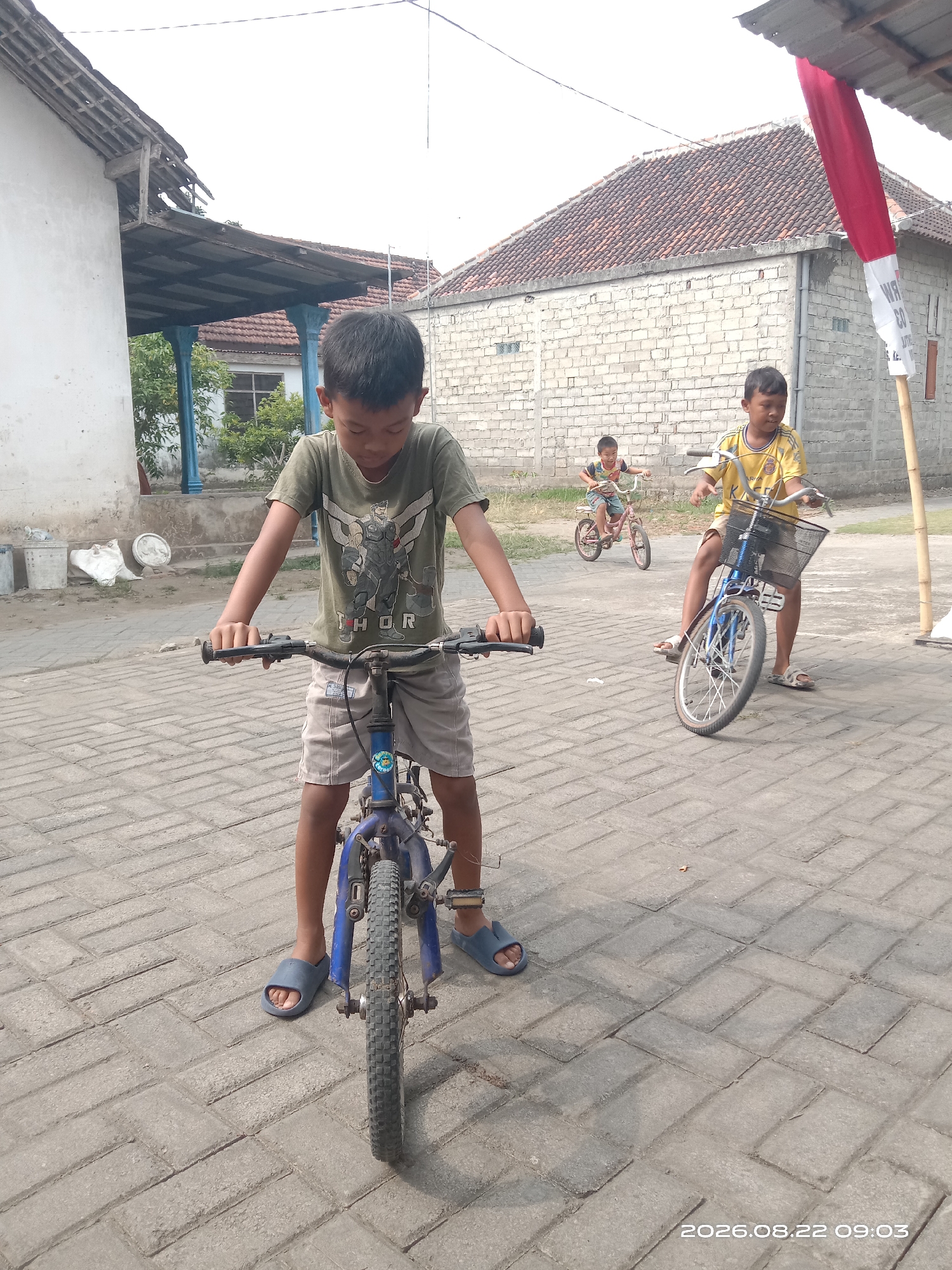 A low-angle, medium shot shows three young boys on bicycles in what appears to be the city of Banyuarang, Indonesia. The boy in the foreground, facing the camera, wears a green t-shirt with a graphic of a character and shorts, and is on a blue bicycle. To his right, a boy in a yellow t-shirt and shorts is on a blue bicycle with a front basket. In the background, a third boy in a colorful t-shirt and shorts is on a pink bicycle. The ground is paved with interlocking bricks. Buildings with tiled roofs are visible behind the children, and a red and white flag is hanging from a pole on the right side of the frame. The sky is overcast. The timestamp 2026.08.22 09:03 is visible in the lower right corner.