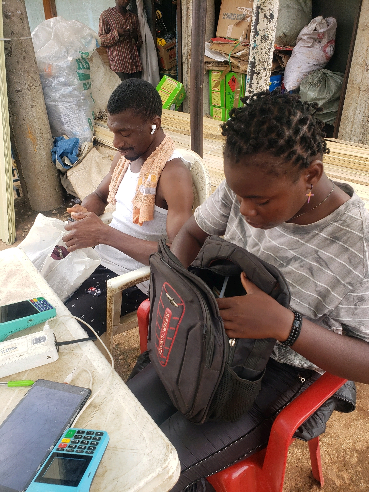 In Sabon Tasha, Nigeria, two individuals are seated at a table. A man in a white tank top with an orange towel draped over his shoulders is looking at his phone while holding a white plastic bag. He is wearing wireless earbuds. Beside him, a woman with braided hair is looking down and appears to be searching inside a black backpack. She is wearing a grey striped t-shirt, a cross pendant necklace, and a beaded bracelet. On the table in front of them are several electronic devices, including two phones, a portable charger, and a blue point-of-sale terminal. The background shows a cluttered area with sacks, boxes, and wooden planks, suggesting an outdoor or semi-outdoor marketplace or storage space.