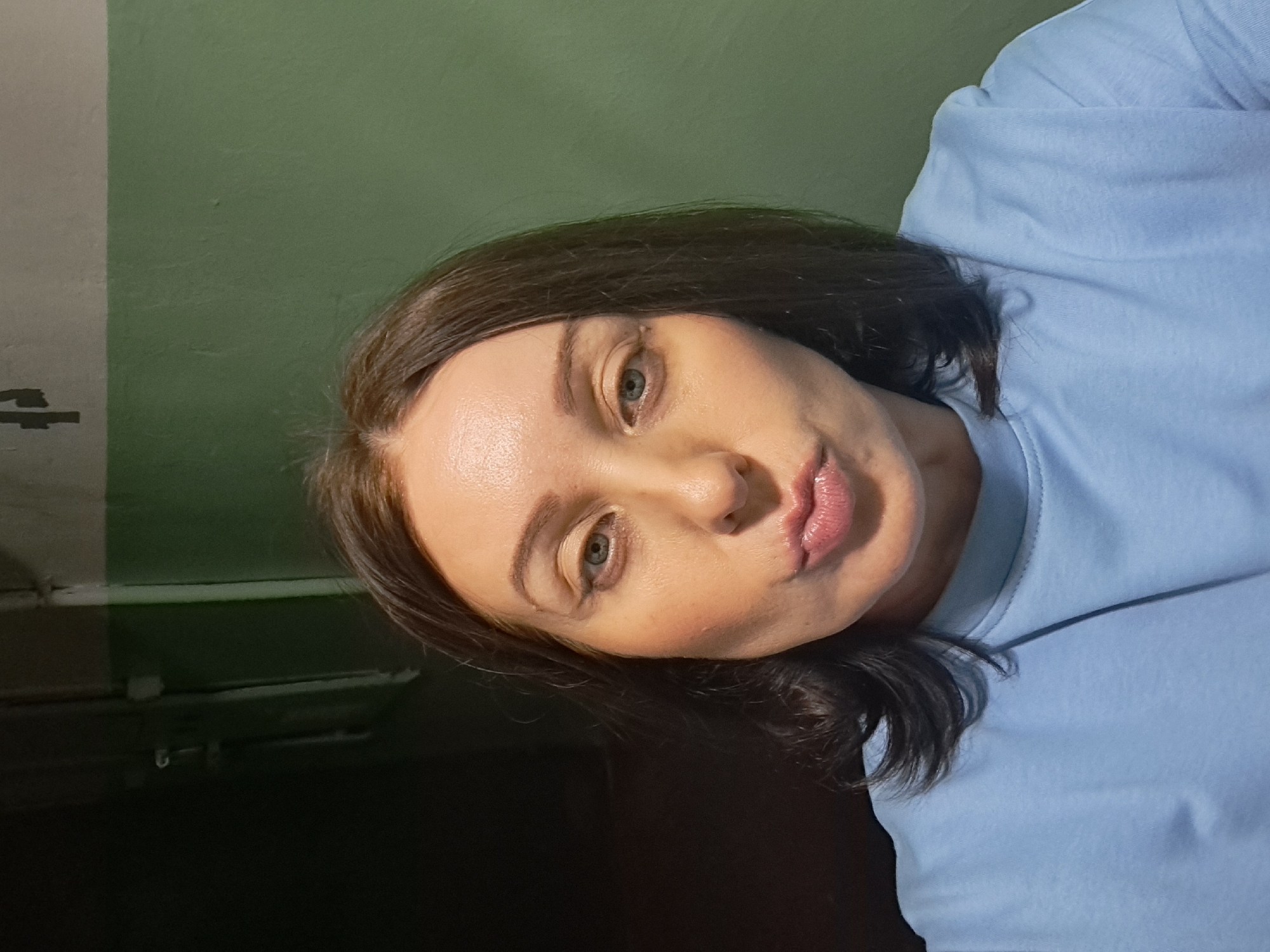 A close-up, waist-up selfie of a woman with medium-length brown hair is shown. She is wearing a light blue long-sleeved shirt. The woman has fair skin, light blue eyes, and is puckering her lips. Her makeup appears to include foundation and lip gloss. The background consists of a dark wall on the left and a green and grey painted wall on the right. The lighting is dim, suggesting an indoor environment. The specific location in Irkutsk, Russia, cannot be determined from the image alone.