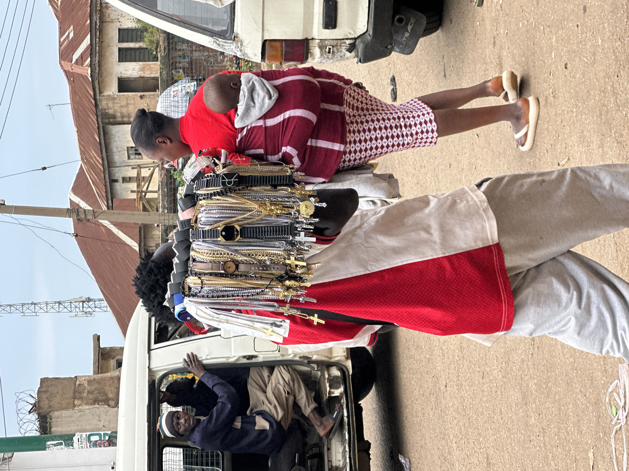 A street scene in Bukuru, Nigeria, captures a vendor walking while carrying an array of necklaces and watches on a display rack. The vendor, wearing a red and white garment and grey sweatpants, has a baby strapped to their back. To the left, a man sits in the open door of a white van, wearing a navy blue sweater and khaki pants. The background features buildings with corrugated metal roofs and overhead power lines, indicative of an urban environment. The ground is paved with dirt and some debris.