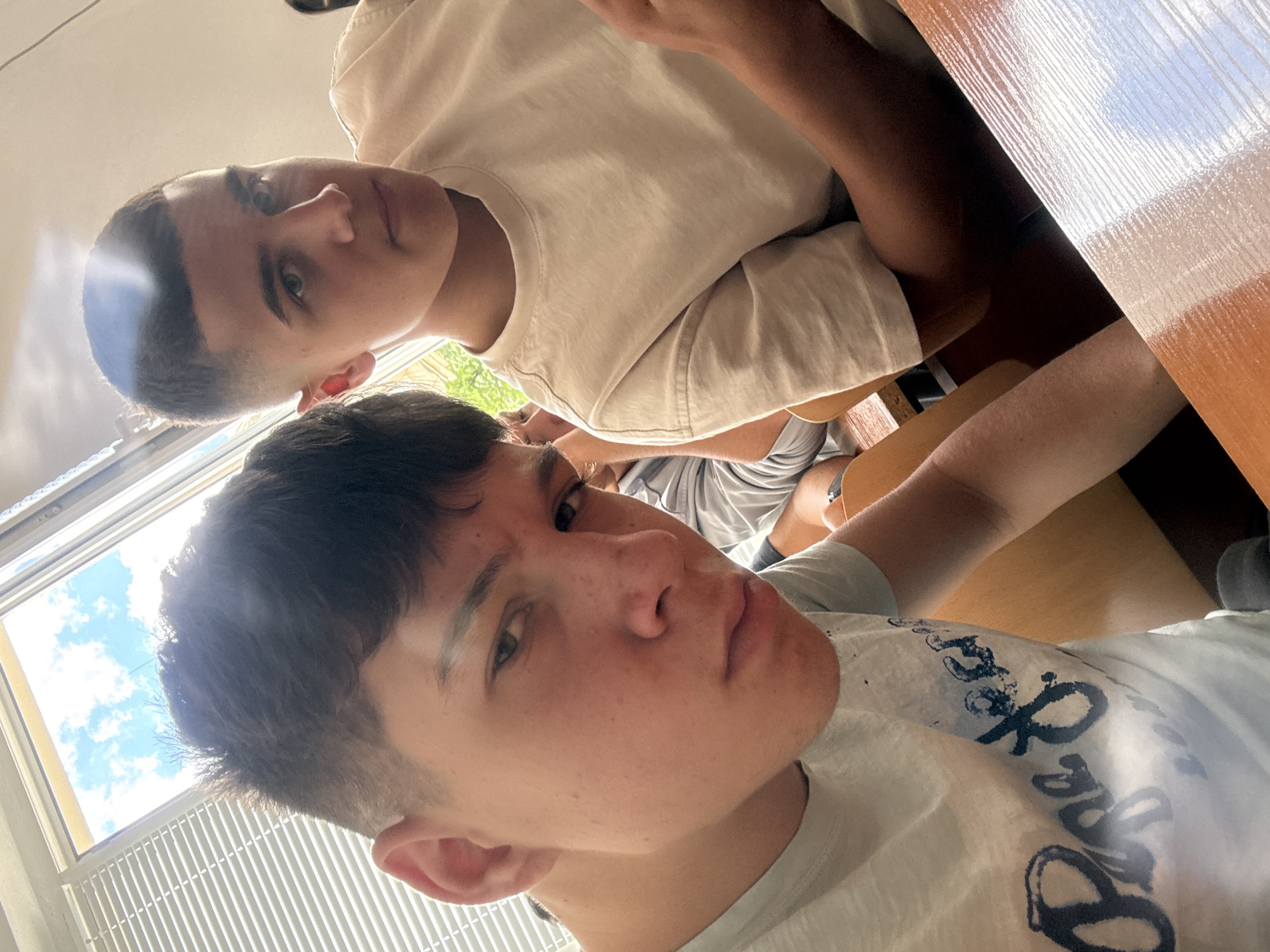 Two young men are sitting at desks in a room with a window. One man is wearing a light-colored t-shirt with a graphic design, while the other is wearing a plain white t-shirt. The room has blinds on the window and a wooden desk. The location cannot be determined from the image.