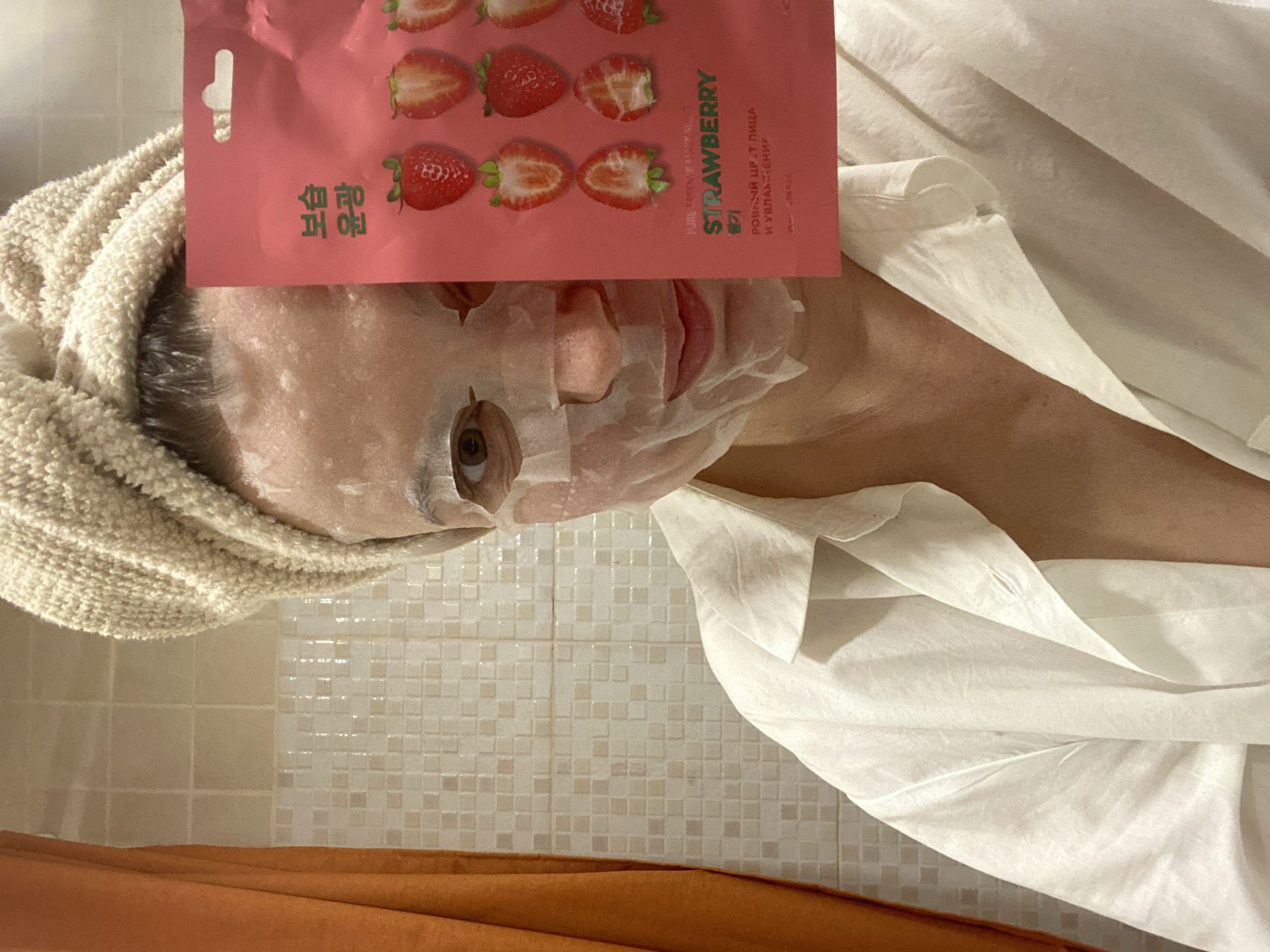 A person lies on their back, wearing a white sheet or garment and a beige towel wrapped around their head. The person has a translucent sheet face mask applied. A pink package with the word "STRAWBERRY" and images of strawberries is positioned above their face. The background shows a light-colored tile wall with small, square tiles, and a glimpse of an orange object at the bottom. The location cannot be verified as Chaikovsky, Russia based on the image.
