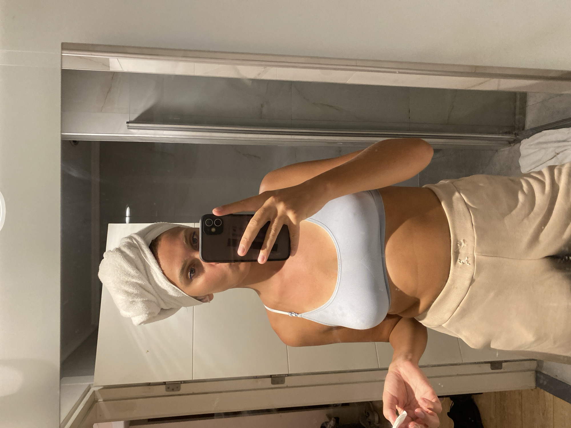 A woman, likely in her 20s, stands in a bathroom, taking a selfie in a mirror. She has a white towel wrapped around her head, covering her hair. She is wearing a light blue sports bra and beige sweatpants. Her left hand is raised, holding a cotton pad. The background shows a shower stall with glass doors and tiled walls. The lighting is ambient, and the overall impression is one of a post-shower grooming or self-care routine. The location is identified as Aksu, Turkey.