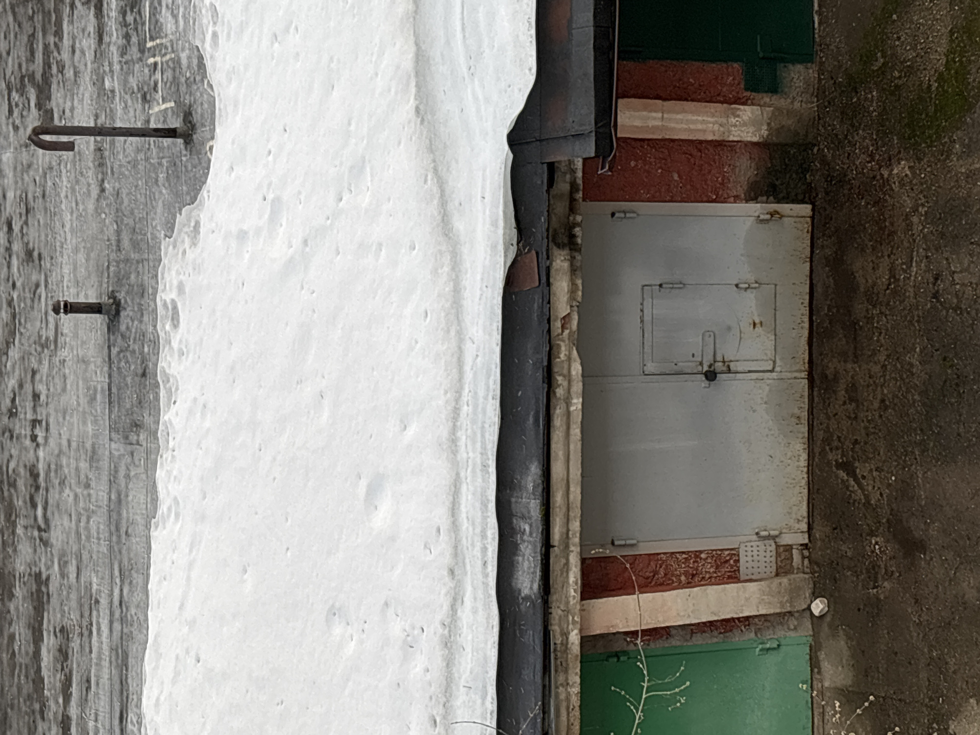 The image depicts a snowy scene outside what appears to be a garage or storage unit, likely located in Perm, Russia. A large snowdrift rests against a gray, wooden wall, with two metal hooks protruding from it. The garage/storage unit is a two-door structure, painted light gray, with a smaller rectangular section on the upper half of one door. It is flanked by sections of red and green paint above and below, and appears to be closed with a padlock. Adjacent to the storage unit is a darker, possibly asphalt, surface.
