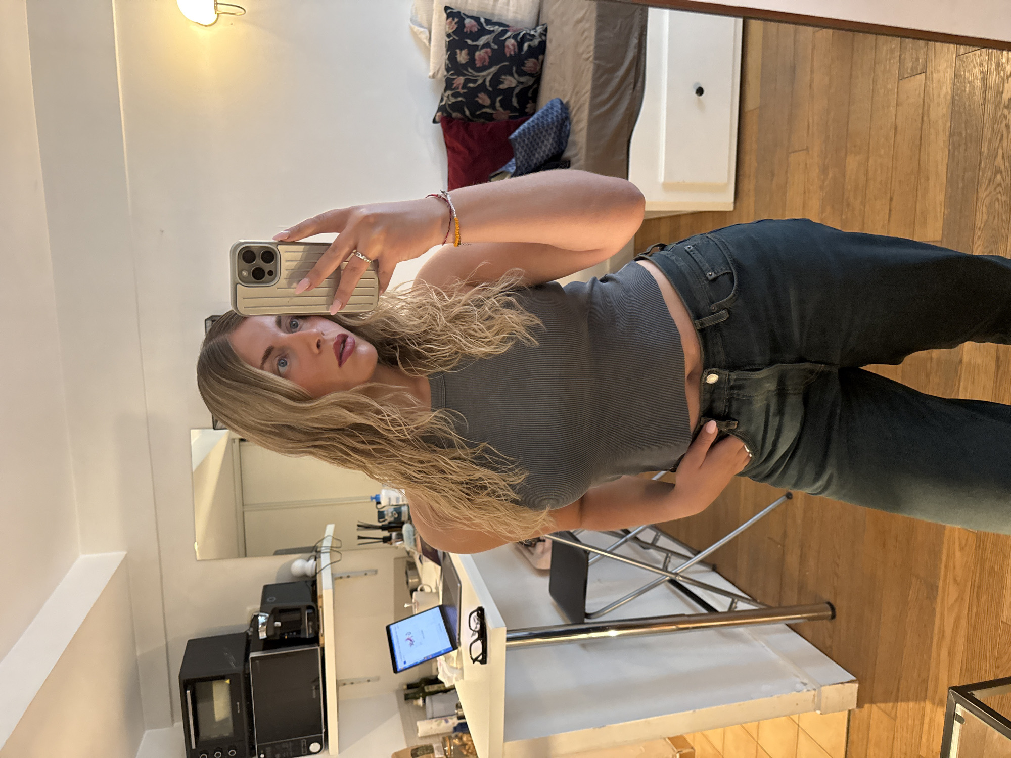 A woman with blonde, curly hair takes a mirror selfie in what appears to be an apartment in Paris, France. She is wearing a grey ribbed tank top and dark wash jeans. Her right hand holds a smartphone, and her left hand is casually placed in her jeans pocket. On her right wrist, she wears two bracelets, and a ring adorns her left hand.
The background reveals a kitchen area with a white countertop, a laptop, a mug, and a pair of glasses on the counter. Above the counter, there are two microwaves stacked on a shelf. To the right of the kitchen counter, a section of a living space is visible, featuring a bed with a brown bedspread and several pillows, one of which has a floral pattern. A white dresser is partially visible on the far right. The flooring is made of light-colored wood planks. The lighting appears to be natural, supplemented by a wall-mounted lamp. The overall scene suggests a domestic setting within an urban environment.