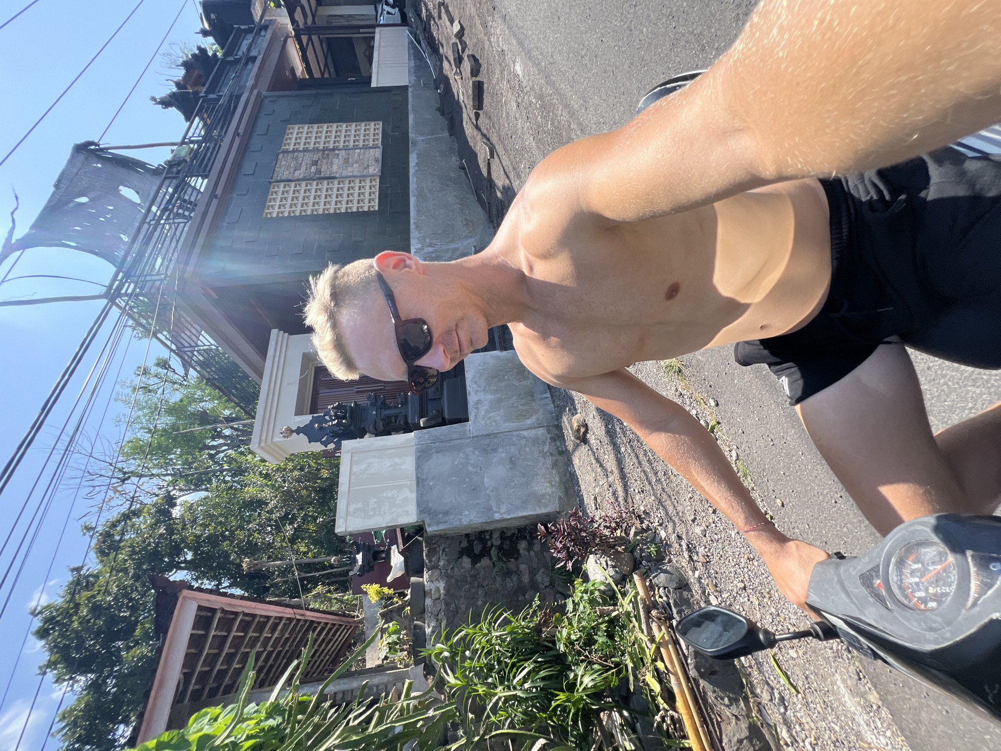 A shirtless man with blond hair and sunglasses rides a scooter on a paved road in Abang, Indonesia. He is wearing black shorts. The scooter is black with a silver and black dashboard. To the left of the road is a stone wall covered in green plants and bamboo. Behind the wall is a pink-roofed building with wooden beams. To the right of the road is a dark green building with patterned windows and a black railing with a ripped tarp overhead. The sky is clear and blue. The lighting suggests it is midday.