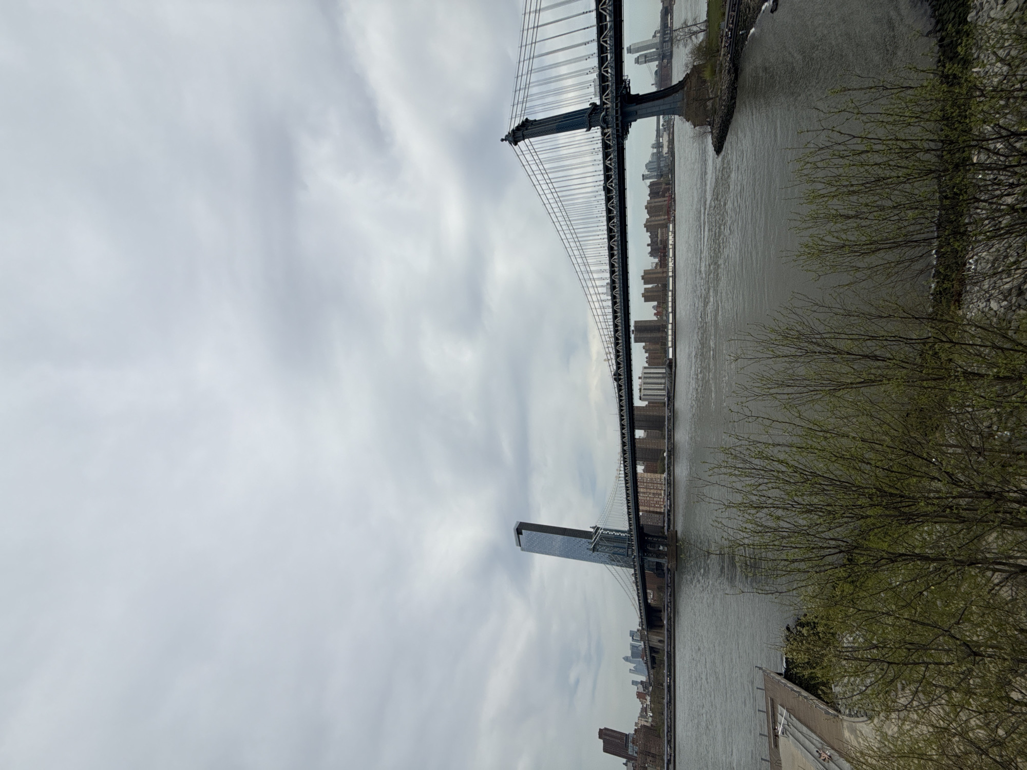 The image depicts the Manhattan Bridge, a suspension bridge that crosses the East River in New York City, connecting Lower Manhattan with Downtown Brooklyn. The bridge's steel cables and towers are prominent features of the image. A body of water runs parallel with the bridge and can be seen to the right. The tops of buildings are visible beyond the bridge. There are trees on the banks to the right. The sky is overcast with clouds.