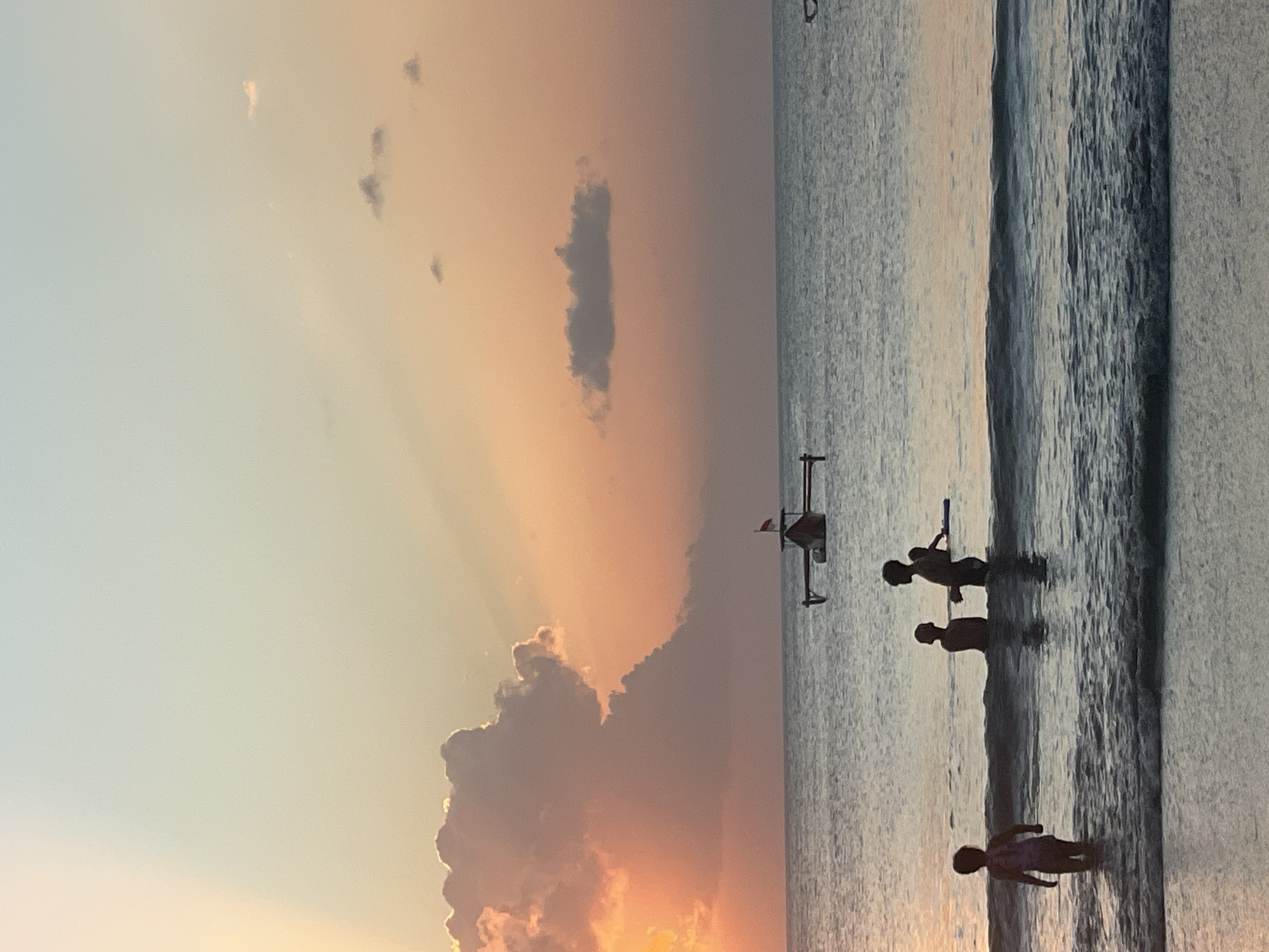 At dusk in Kuta Selatan, Indonesia, several people are silhouetted in the shallow ocean water. In the foreground, a child stands in the waves facing the ocean, with a faint outline of a bathing suit. Further back, two figures, likely children, are standing together in the water. To their right, a larger silhouetted figure, possibly an adult, stands with their back to the viewer. Another figure is partially visible behind them, and a long, blue floatation device rests in the water. In the mid-ground, a traditional Indonesian fishing boat with an outrigger, adorned with a red flag, floats on the calm sea. The sky above is a gradient of soft colors, transitioning from a pale blue at the top to warm hues of orange and pink near the horizon, illuminated by the setting sun. Large, voluminous clouds are present, with one particularly prominent cloud formation to the left, catching the last rays of sunlight. The ocean surface is calm with gentle waves breaking near the shore.