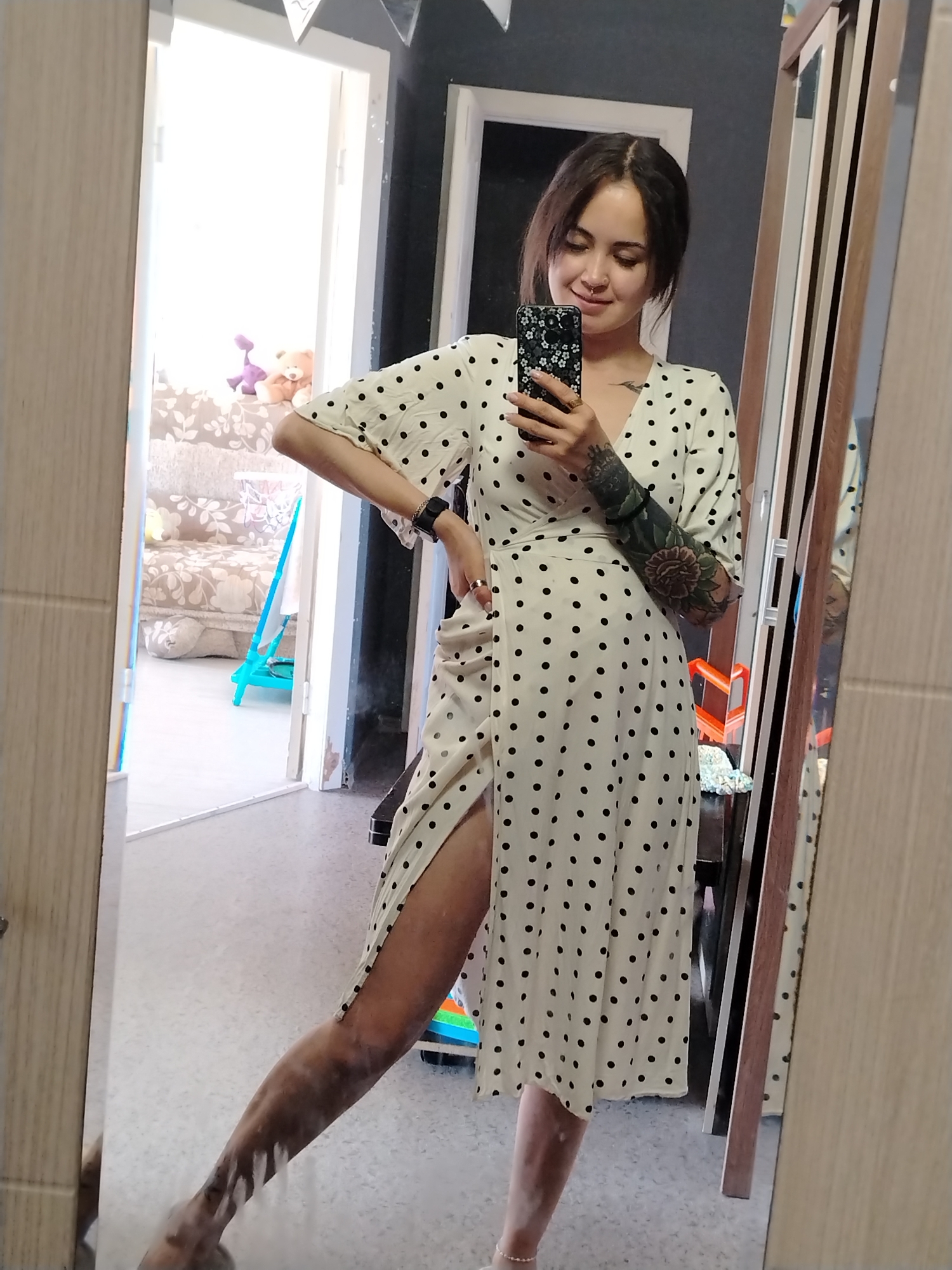 In Noviy, Russia, a young woman with dark hair, wearing a white and black polka dot dress and a nose ring, stands in front of a mirror. She is holding a smartphone with a floral case in her right hand, taking a selfie. Her left arm is adorned with tattoos, and she wears rings on her fingers and a watch on her left wrist. The mirror reflects a room with an open doorway leading to another area where a sofa and a blue toy are visible. The woman's right leg is extended forward, with a high slit in her dress revealing her thigh.
