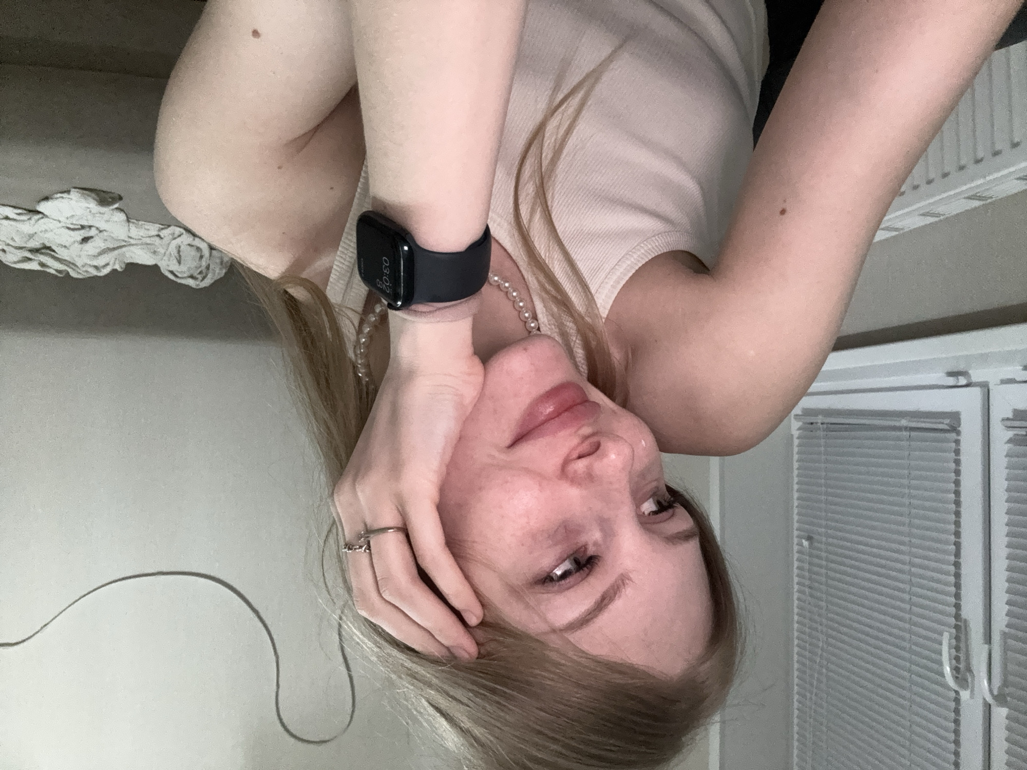 A young woman with fair skin and blonde hair is leaning her head on her hand, looking to the left of the frame with a gentle smile. She is wearing a light-colored tank top and a pearl necklace. On her left wrist, she wears a dark smartwatch. The background shows a window with blinds, a radiator, and a couch with some clothes on it. The lighting is soft and diffused. The scene appears to be an indoor residential setting, possibly an apartment or house, with no specific indicators placing it within the city of Perm, Russia.