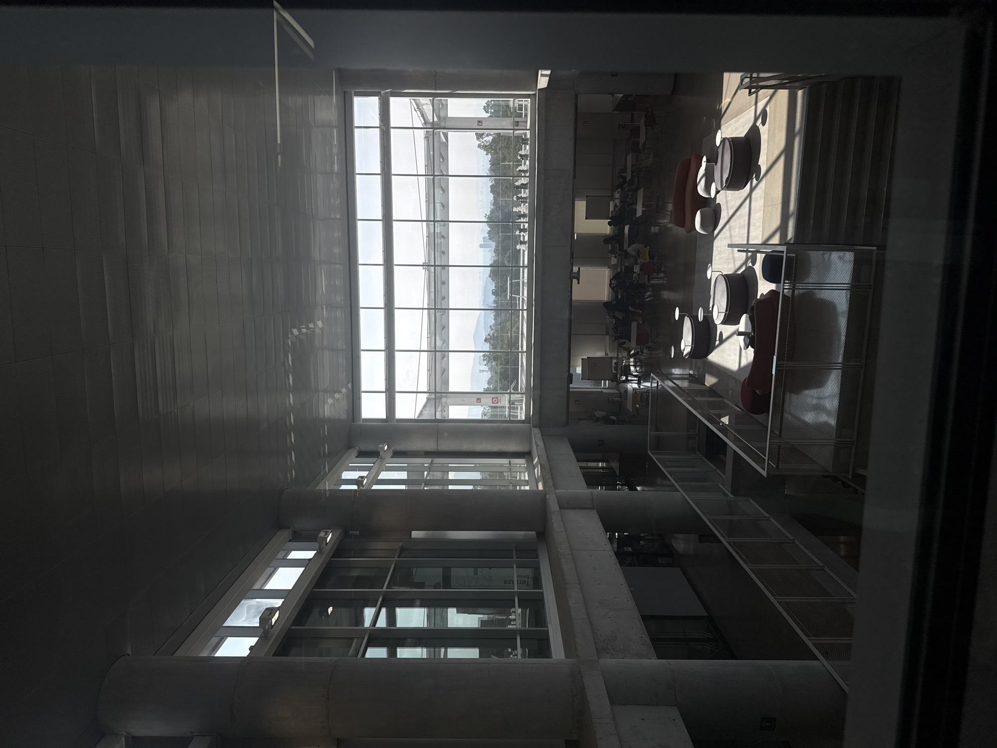 The media file presents an interior view of a modern, multi-level building, likely a university or commercial complex, overlooking an urban and natural landscape. The image is taken from an elevated position, looking down into a spacious common area.
**Subjects:**
* **People:** Several individuals are seated at tables in the lower right portion of the frame (when rotated to landscape). They appear to be casually dressed, engaged in activities such as dining, studying, or conversing. Their faces are not clearly discernible due to distance and lighting.
* **Objects:** The interior features a variety of furniture, including square and round tables, alongside both standard and lounge-style chairs in colors like red/orange, grey, and white. Prominent architectural elements include large concrete pillars and beams, extensive floor-to-ceiling windows with metal framing, and metal mesh railings and structures. Wall-mounted and recessed light fixtures are also visible.
* **Scene:** The scene depicts a bright, open-plan indoor space, possibly a cafeteria, lounge, or study area, characterized by its contemporary design and ample natural light.
* **Setting:** The setting is an interior space in a modern building in Mexico City. Through the large windows, an outdoor view reveals trees, a distant city skyline with various buildings, and mountains or hills in the far background.
**Activity or event taking place:**
People are observed occupying the tables and chairs, suggesting a regular daytime activity such as eating, working, or socializing in a public or semi-public space. There is no specific event apparent.
**Notable details:**
The time of day is clearly daytime, evidenced by the bright sunlight streaming through the windows, casting long, sharp shadows across the floor and furniture. This also indicates clear weather conditions. The distant cityscape and mountain range visible through the windows align with the geographical context of Mexico City. The overall mood appears calm and relaxed. Faint text "Terrazas" and "Terrace" is visible on one of the window panes, suggesting an accessible outdoor area adjacent to the interior space.