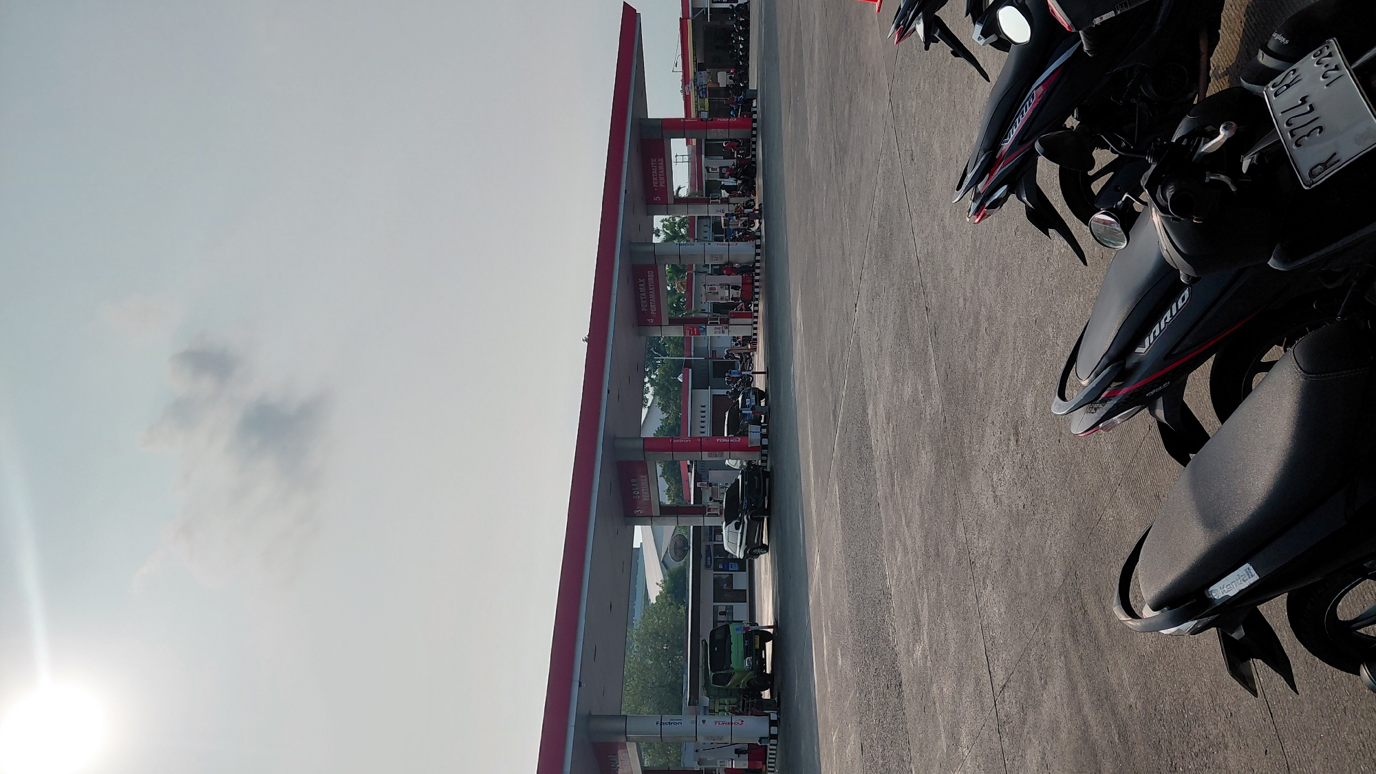 The image shows a gas station with several vehicles, including cars and motorcycles. A green truck is parked on the left side of the gas station, and a black SUV is parked in the middle. Several motorcycles are parked in front of the gas station. The gas station has a red roof and white pillars. There are also some trees and buildings in the background. The sky is clear and the sun is shining. The scene appears to be in Karanglewas, Indonesia.