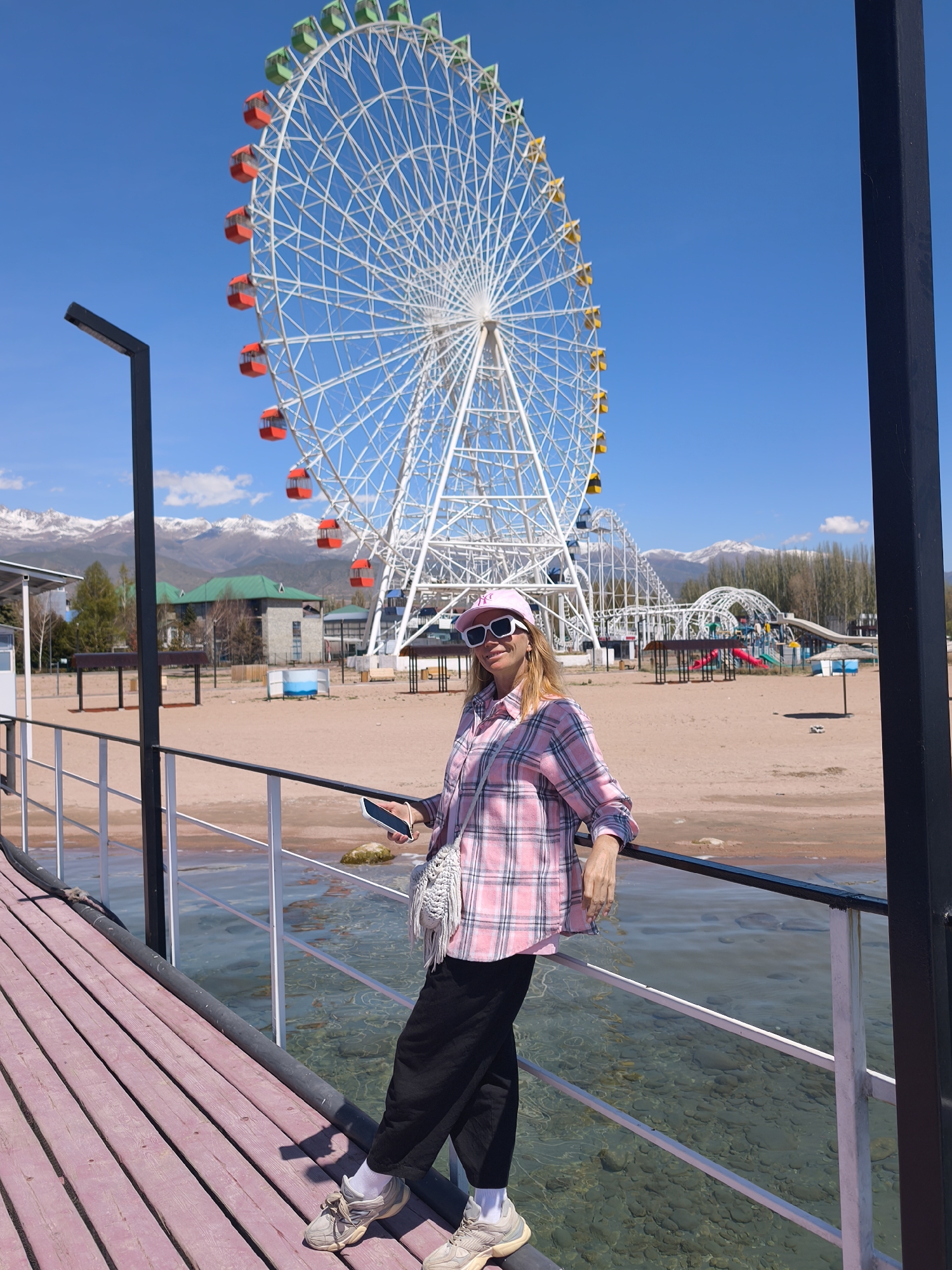A medium shot captures a woman standing on a pier with a large Ferris wheel in the background. The woman is leaning against the pier's railing, holding a smartphone in her right hand, and wearing a pink plaid shirt, dark pants, and a pink baseball cap with sunglasses. She has a white crossbody bag on her left side. The Ferris wheel, white with colored gondolas, dominates the backdrop under a clear blue sky. Mountains are visible in the far background. The pier has a pink-toned wooden deck with black and white railings, extending over clear water that reveals underwater stones. Further in the background, there is a sandy beach with structures including buildings, trees, and a playground. The scene is situated in Bosteri, Kyrgyzstan.