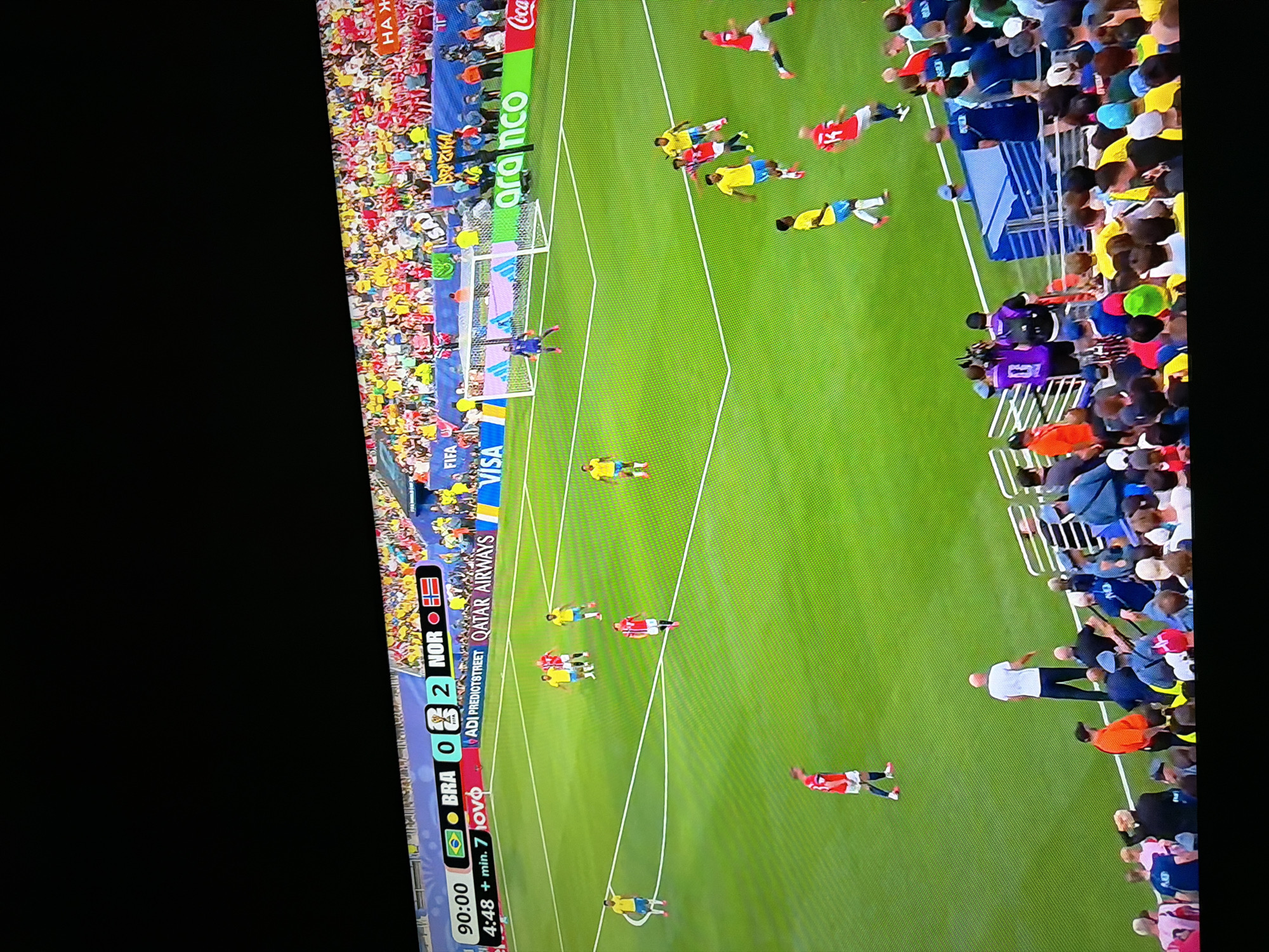 This is a photograph taken of a television screen displaying a soccer match. The screen shows a score overlay at the top indicating "BRA 0 - 2 NOR" at "90:00" with "4:48 + min. 7 ovov". The match appears to be in its final moments, with a soccer ball visible on the green field. There are several players in yellow and red uniforms on the field, as well as a goalkeeper near the goal. Spectators are visible in the stadium seating area behind the field, many wearing yellow and green attire, likely supporting Brazil. The environment is a brightly lit soccer stadium with advertisements and sponsor logos visible along the sidelines. The image quality suggests it was taken at an angle and possibly with flash, resulting in glare and distortion. There is no discernible connection to Ognianovo, Bulgaria in this image.