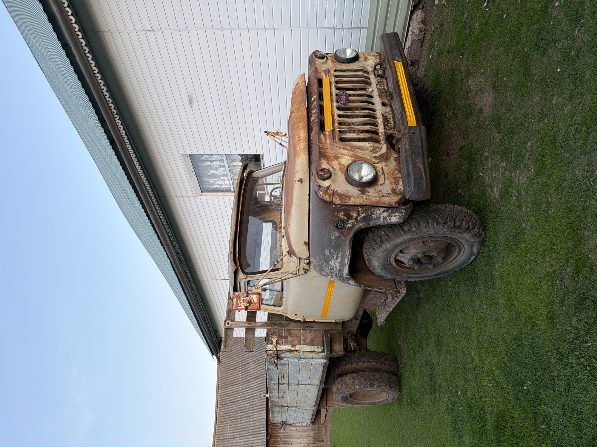 The image shows a severely rusted GAZ-53 truck parked near a building with a white siding. The truck is heavily weathered and appears to be non-operational. The surrounding ground is covered in grass. The building appears to be of utilitarian construction. Based on visual cues and the provided location, the environment seems to be a rural or semi-rural setting in Кадряково, Russia. There are no visible people or interactions in the scene. The focus is solely on the stationary, dilapidated truck within its environmental context.