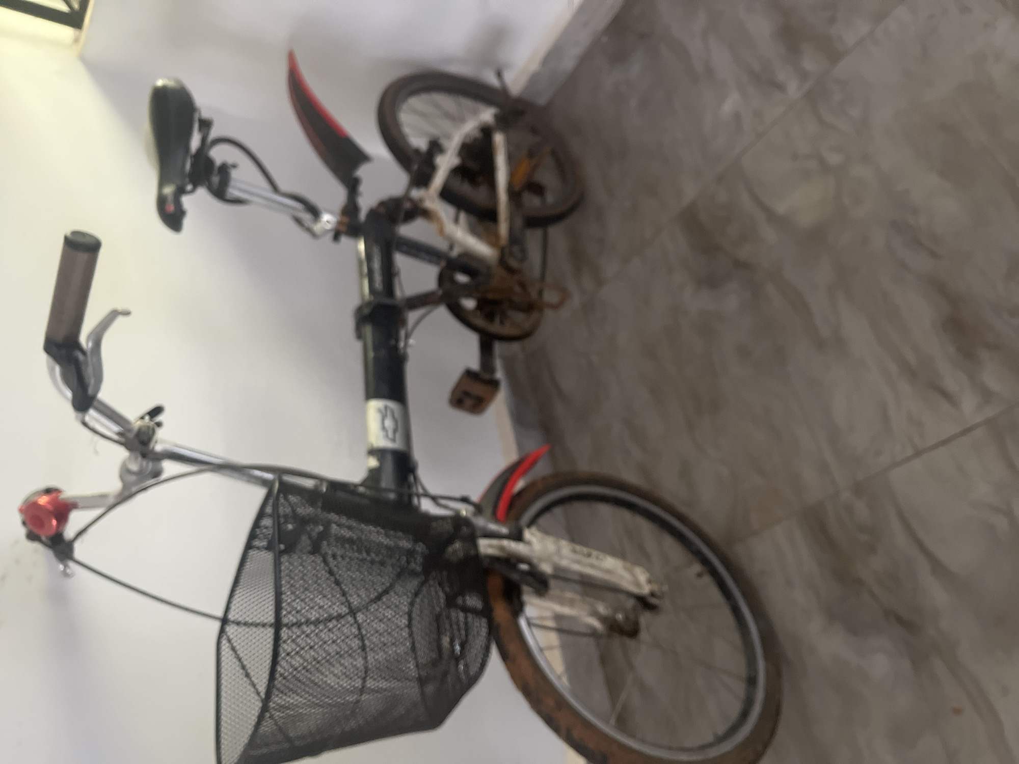 This is a full-shot, medium-angle, eye-level, blurry image of a black folding bicycle with a large black wire basket mounted on the handlebars. The bicycle is standing upright on a tiled floor inside a building, likely a home or storage area. The bicycle is positioned at an angle, with the front wheel closer to the camera and facing left. The bicycle has a black frame, a black seat, and a red bell on the handlebars. The front wheel has a muddy tire and a white fork. The rear wheel is partially visible. The background is a plain white wall. The image quality is poor, with low resolution and a lack of focus, making it difficult to discern finer details. There is no indication of the location being in Jos, Nigeria, as no specific landmarks or context are provided.
