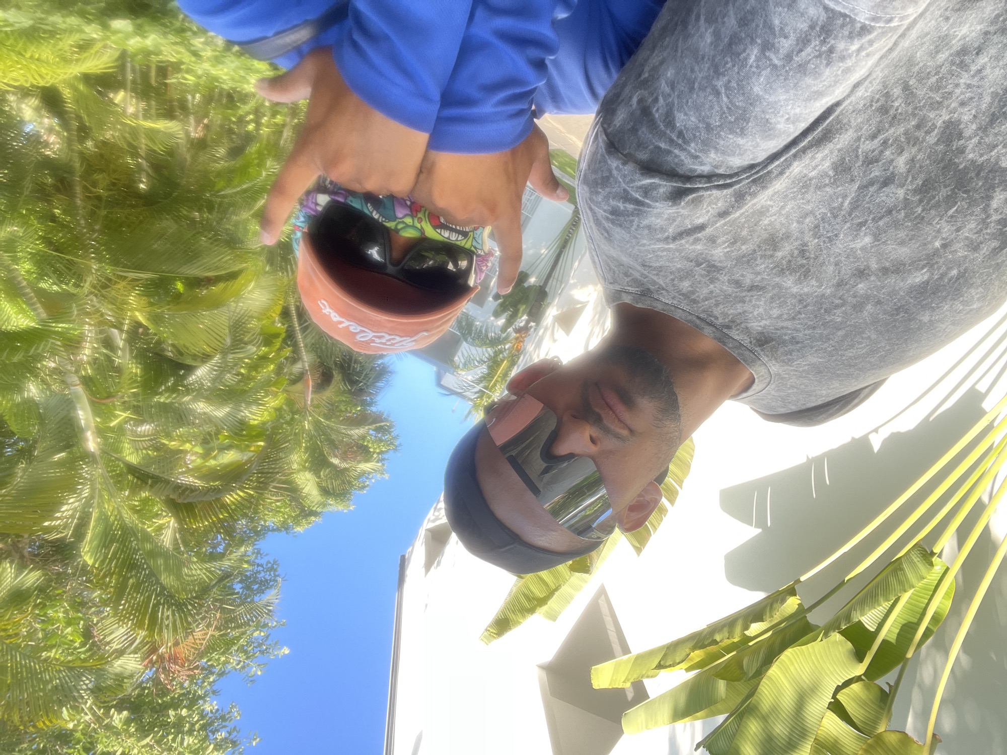 The image depicts two individuals in Playa del Carmen, Mexico, likely taking a selfie. One person is wearing a blue long-sleeved shirt, a Titleist golf hat, and sunglasses, while the other is wearing a gray t-shirt and sunglasses with a black headband. They are standing outdoors, with tropical vegetation including palm trees and banana plants in the background. A white building with louvers is partially visible. The bright blue sky suggests a sunny day. The photograph is taken from a low angle, looking upwards.
