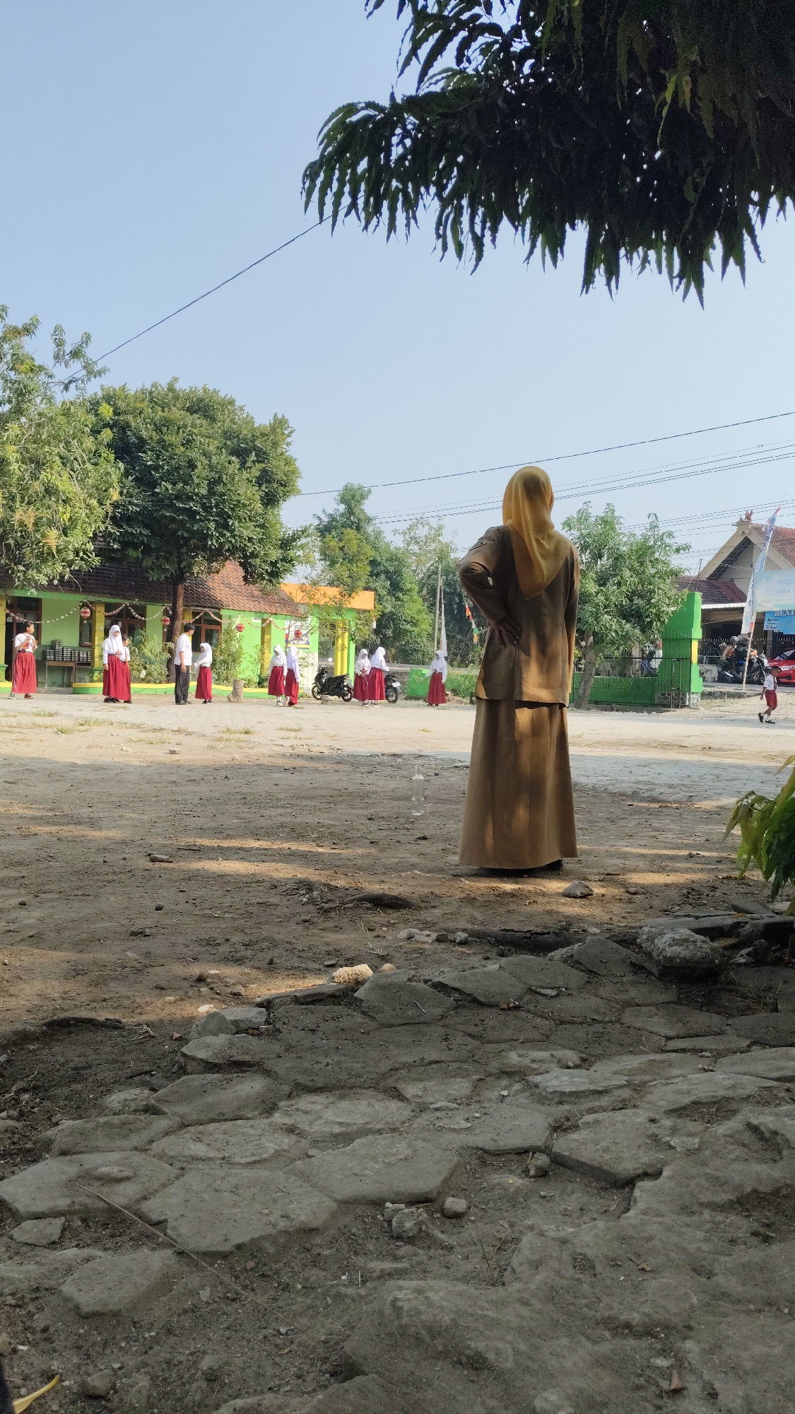 A wide-angle, eye-level shot captures an outdoor scene at what appears to be a school in Banjarsari, Indonesia. The foreground is dominated by a rough, cobbled ground surface, with scattered dirt and debris. In the midground, a figure, likely a teacher, stands with their back to the camera, wearing a long brown skirt and a tan jacket with a yellow hijab. The teacher's hands are on their hips, suggesting a watchful stance.
Further in the midground, a group of schoolchildren, all wearing red skirts and white tops, are scattered across a dusty courtyard. Some are standing in groups, while others appear to be walking. A few motorcycles are parked nearby, one of which is partially obscured by a group of students. The background features a low-rise, light green building with a tiled roof, possibly a classroom or school administrative building. Trees with lush green foliage surround the buildings, and a bright blue sky fills the upper portion of the image. The overall impression is a typical school day in a sunny, rural or semi-rural Indonesian setting.