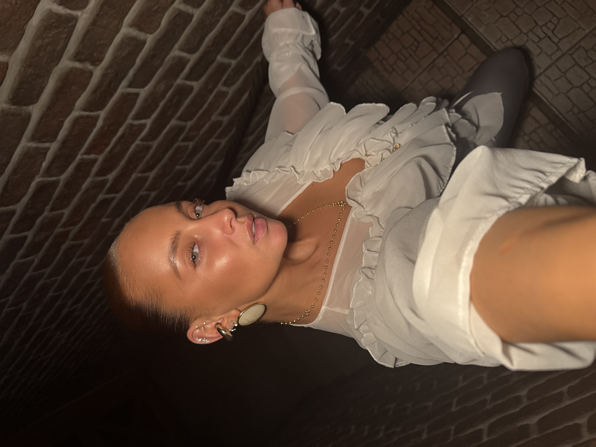 A woman, whose skin is illuminated by bright light, is looking towards the camera. She has her hair pulled back, and is wearing large gold earrings and a white ruffled blouse. She is standing in what appears to be an indoor area with a brick wall behind her and a patterned floor. The location is not specified in the city of Chaikovsky, Russia.
