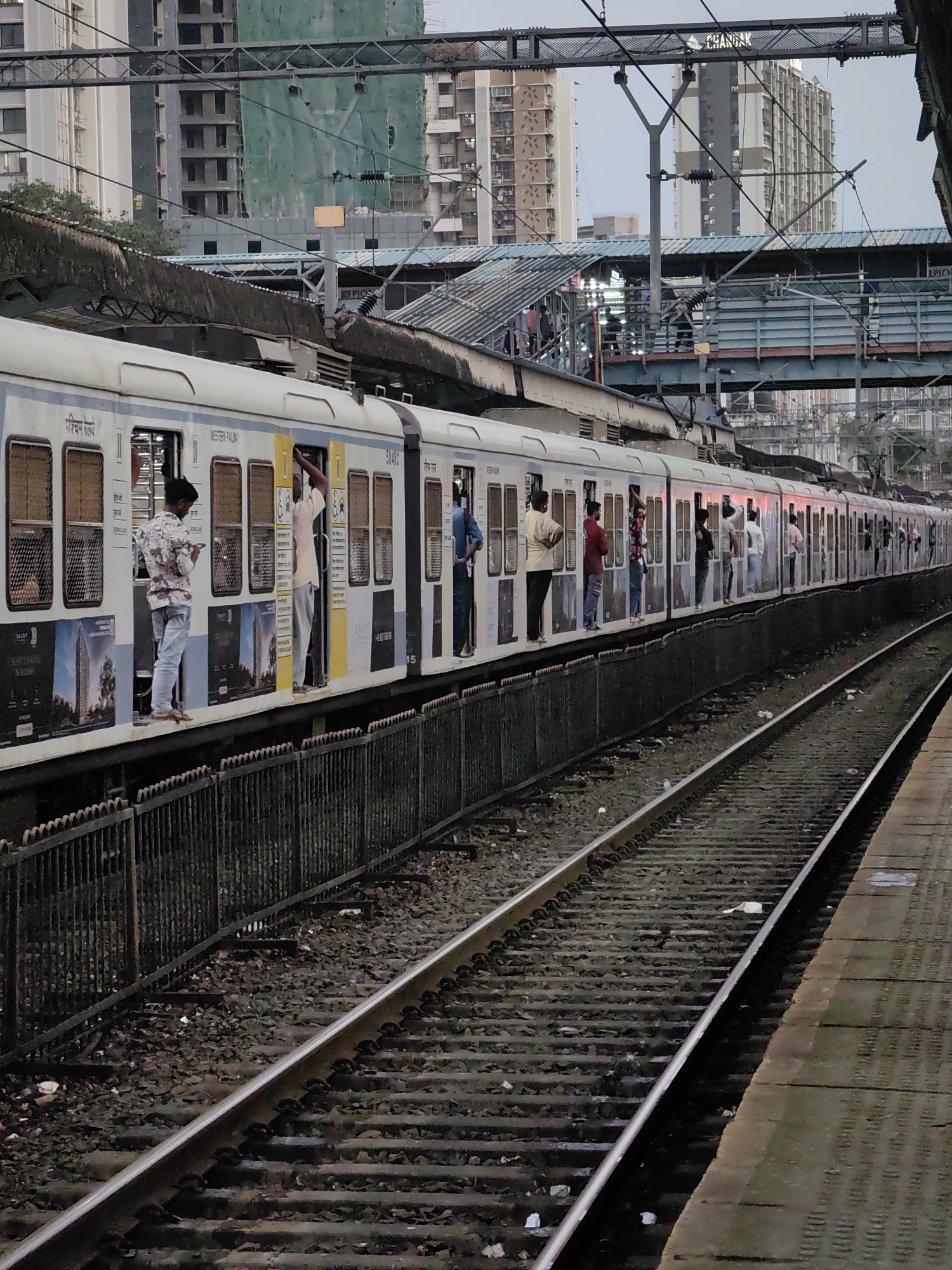 A long, white suburban train with blue and yellow accents is stopped at a train station in Mira Bhayandar, India. The train has numerous open doors and windows, with people standing in, leaning out of, and hanging onto the sides of the train cars. The platform is adjacent to the train tracks, and a dark metal fence runs along the edge of the platform. In the background, there are elevated train tracks, a pedestrian overpass, and several tall buildings under construction, one covered in green netting. The sky is overcast.