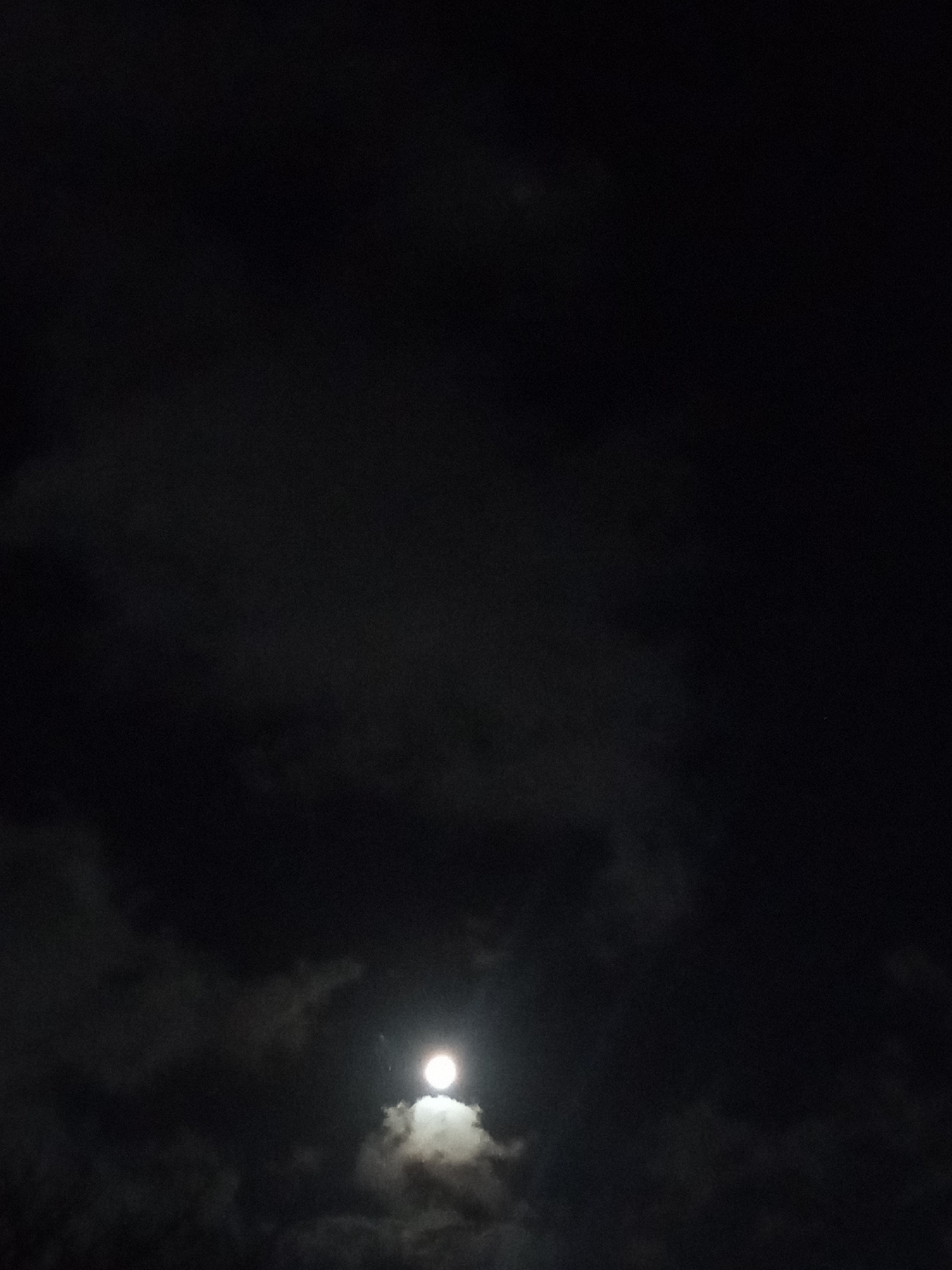 The image shows a night sky with the moon visible through a layer of clouds. The moon is positioned towards the lower center of the frame, and appears bright against the dark sky and clouds. The overall environment suggests a dark night, possibly in an outdoor setting where the moon is the primary source of illumination. There is no other information in the image to confirm its location in Juan Griego, Venezuela.