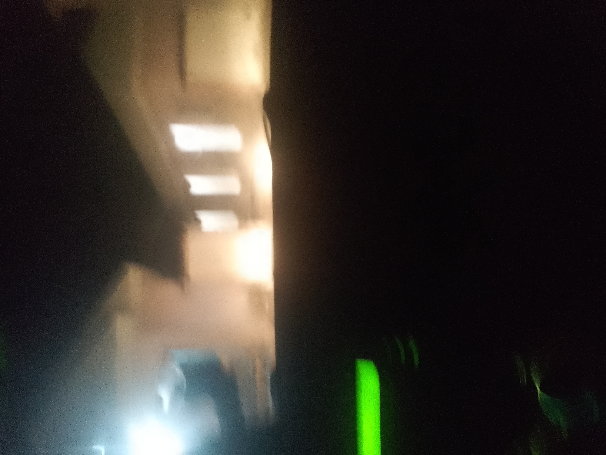 The image depicts a dark, indoor environment with artificial lighting. In the left portion of the image, there are several bright rectangular light sources, suggesting windows or illuminated panels. The overall lighting is dim, with a yellowish cast, indicating artificial illumination. The right side of the image is predominantly black, with some faint green light streaks visible towards the bottom right, possibly from electronic devices or ambient light. The scene appears to be a dimly lit room or corridor. Without further information or clearer visual cues, it is impossible to pinpoint the specific location in Jalingo, Nigeria.