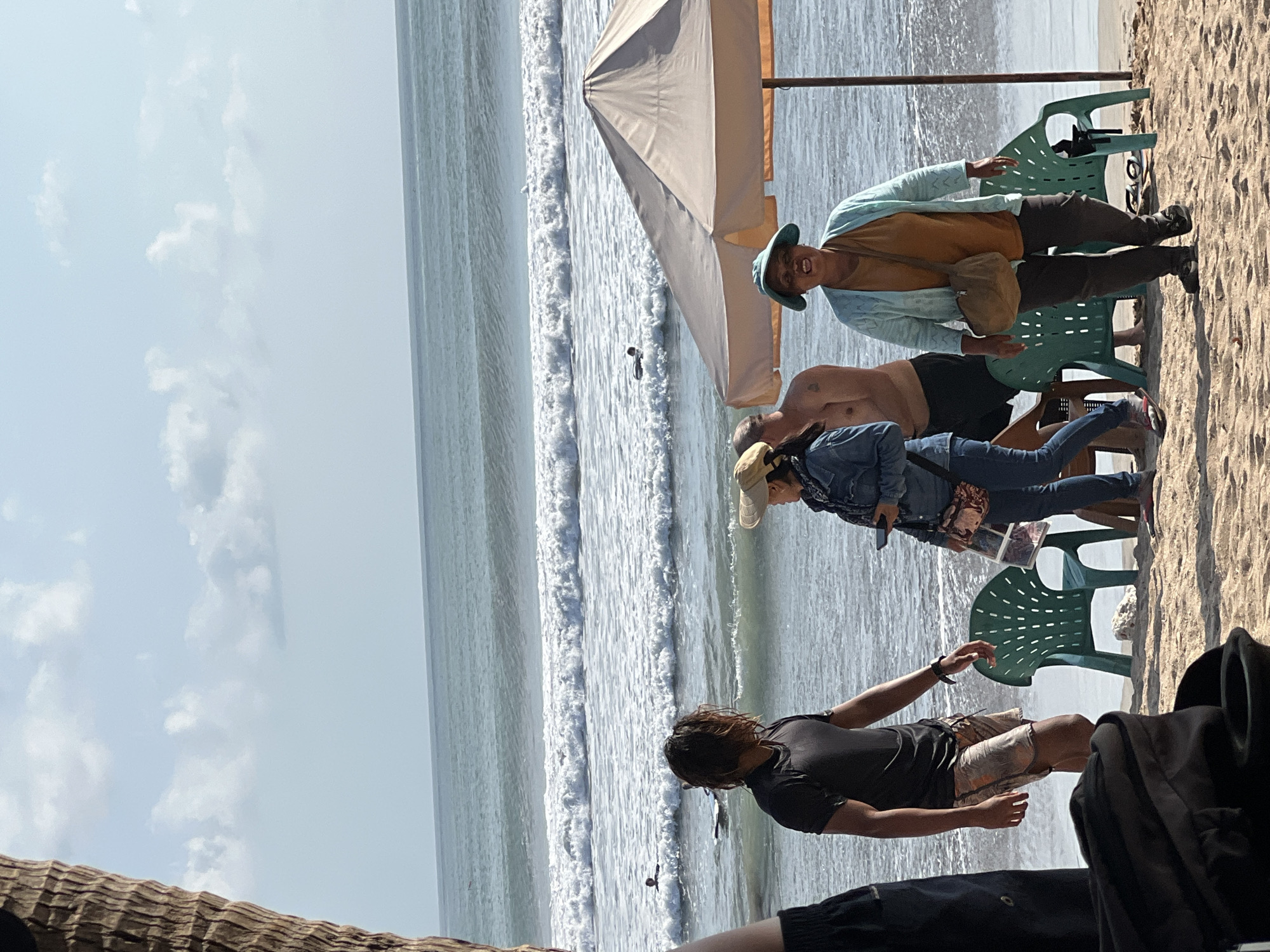 In Kuta, Indonesia, on a sandy beach, several individuals are present near the ocean. A man with long, dark hair, wearing a black t-shirt and patterned shorts, stands looking towards the water. To his right, a woman in a denim jacket and light-colored hat is visible, holding a phone. Behind her, a shirtless man stands partially obscured. In the foreground, a woman wearing a wide-brimmed blue hat, an orange shirt, and a light blue cardigan, with a crossbody bag, is walking towards the viewer. Two green plastic chairs are positioned on the sand near the water's edge. An umbrella provides shade over a section of the beach. The ocean displays moderate waves, and a few surfers are in the water. The sky is partly cloudy.