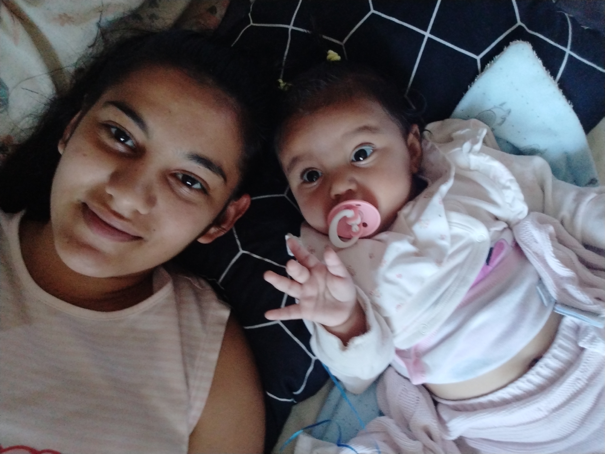 The image shows two individuals, presumably a young woman and a baby, lying on a bed. The woman is positioned on the left side of the frame and is looking at the camera with a slight smile. She has dark hair and is wearing a light-colored tank top. To the right of the woman is the baby, lying on its back. The baby has a pacifier in its mouth and is wearing a light-colored outfit with a blanket. The bedding consists of a pillow with a geometric pattern and a light blue blanket. The location in the city of Unknown, Unknown is not provided.