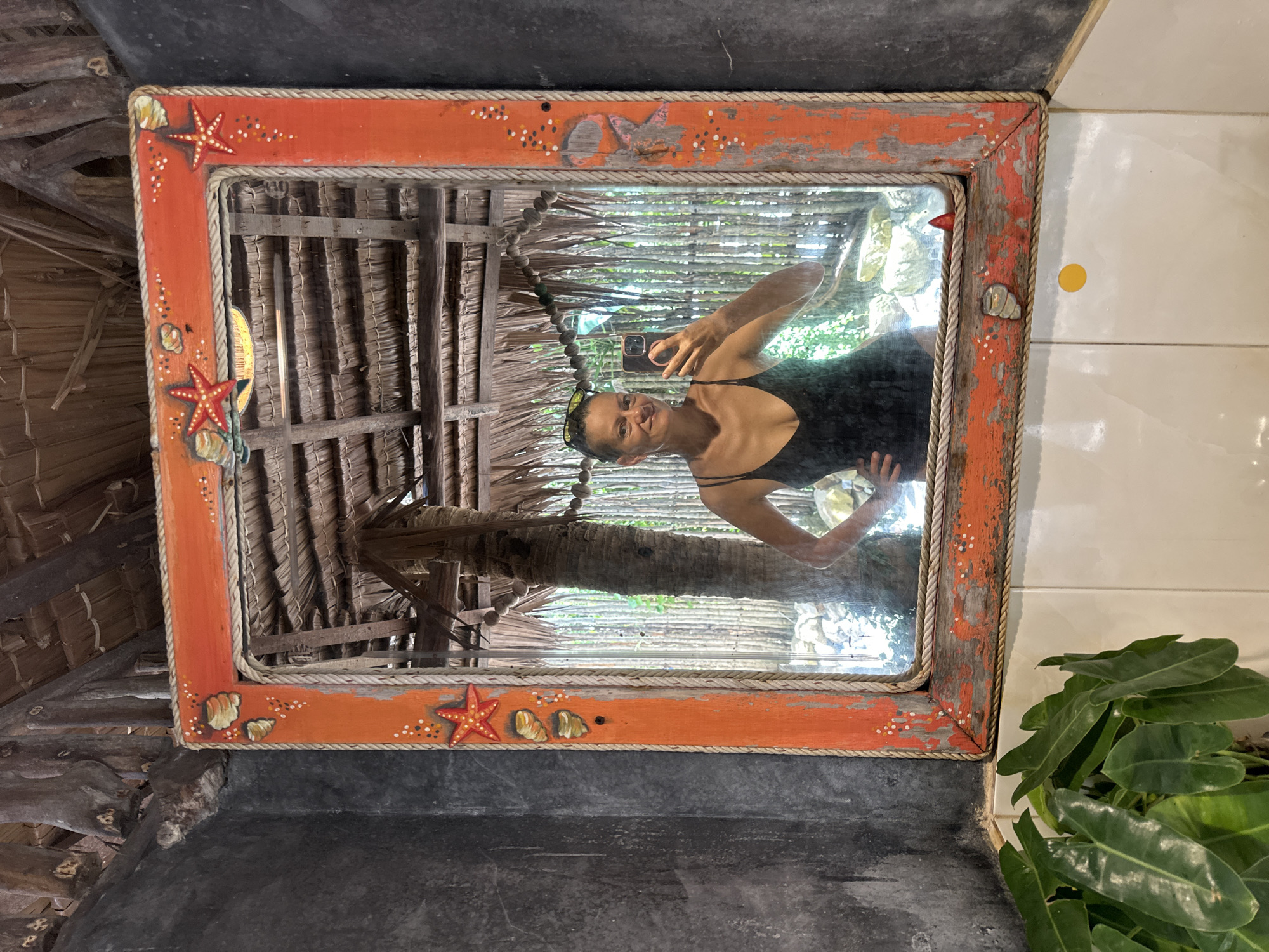 A woman is taking a mirror selfie in a rustic bathroom. The mirror has an orange, weathered frame decorated with shells and starfish. The background reflected in the mirror shows a thatched roof structure and palm trees, suggesting a tropical outdoor setting, possibly in a resort or beachside establishment in Koh Phangan, Thailand. The woman is wearing a black spaghetti strap top and sunglasses perched on her head. The bathroom wall to the left of the mirror is dark and textured, while the wall below the mirror is tiled in white. A large leafy green plant is visible in the lower left corner.