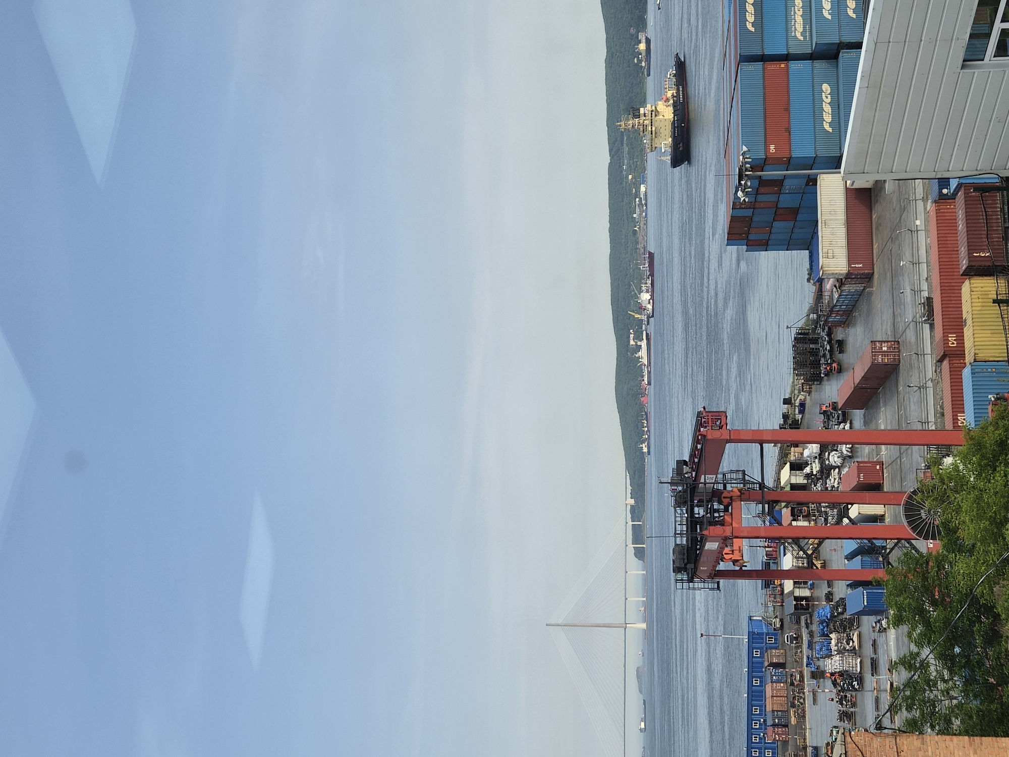 The image captures a view of the commercial port of Vladivostok, Russia, with the Golden Bridge visible in the background. The foreground features a red gantry crane positioned over a bustling cargo yard filled with stacked shipping containers in various colors, primarily blue and red. Numerous other containers are arranged on the ground, alongside vehicles and equipment typical of a port facility. Several smaller vessels, including tugboats and cargo ships, are present in the bay, navigating the waters in front of a tree-covered hillside. The sky is clear and pale blue, with some subtle reflections visible on a transparent surface in the upper portion of the image, suggesting the photo was taken through a window. The overall scene depicts a working port environment under daylight conditions.