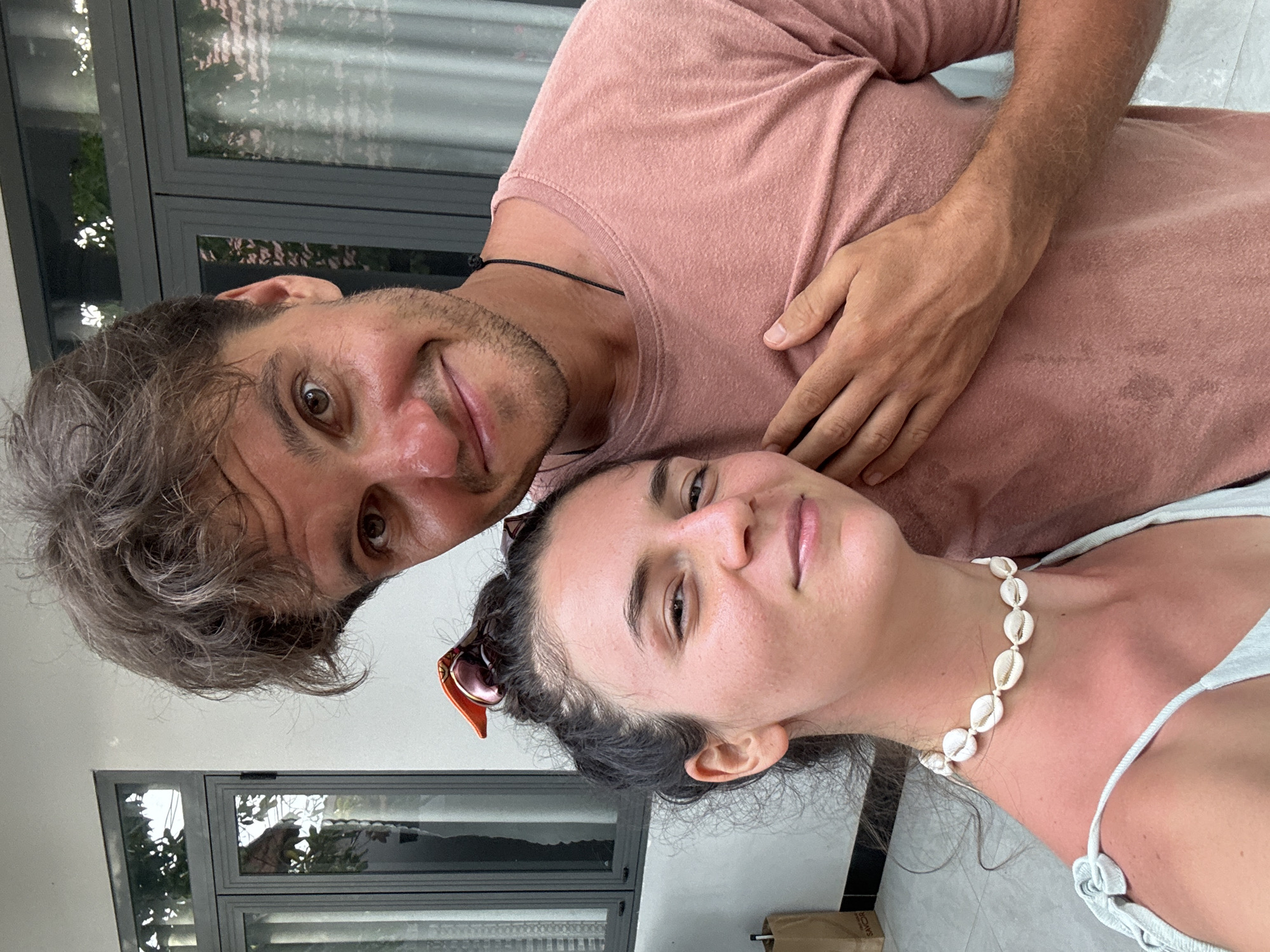 A medium shot shows a man and a woman posed closely together. The man, on the left, has curly, light brown hair with streaks of grey. He is wearing a pink t-shirt. The woman, to the right, has dark hair pulled back and sunglasses perched on top of her head. She is wearing a white tank top and a necklace made of seashells. They are in Mũi Né, Vietnam, standing in front of a building with grey window frames. A reflection can be seen in the window glass.