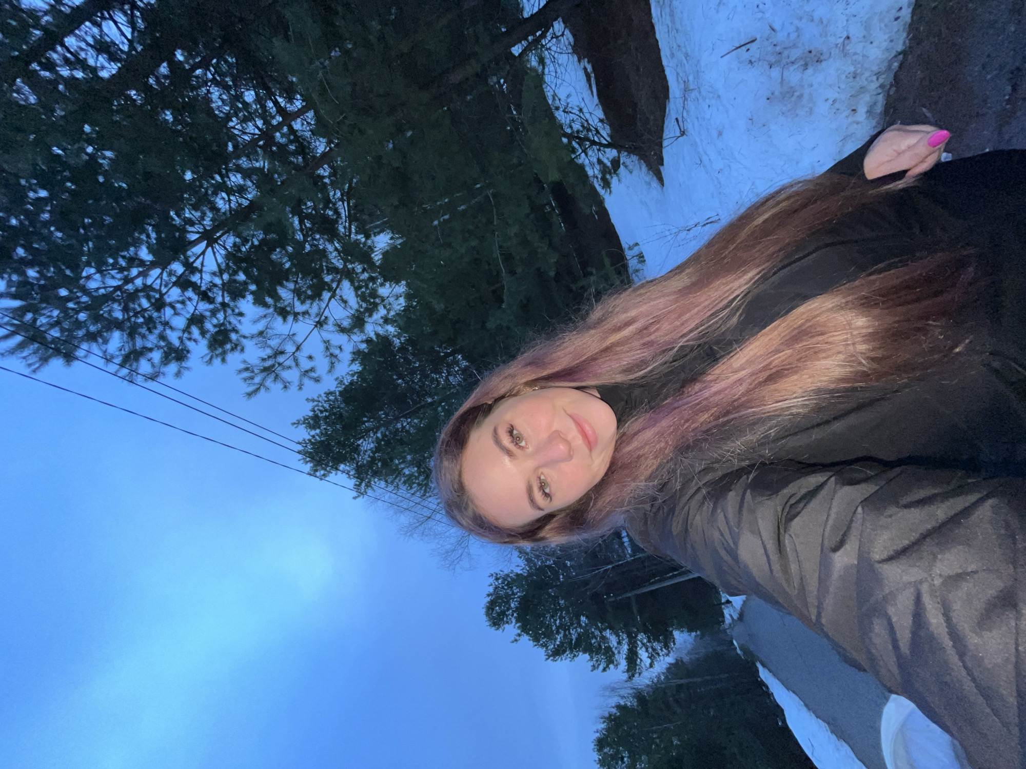 A woman is taking a selfie outdoors. She has long brown hair with hints of purple. She is wearing a black turtleneck and a dark jacket. Her fingernails on one hand are painted pink. In the background, there are trees, a blue sky, power lines, and patches of snow. It is dusk or dawn. Location: Chaikovsky, Russia.