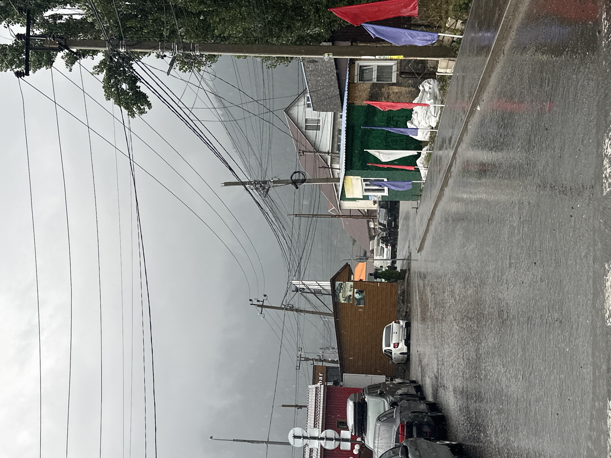 The image depicts a street scene in the city of Arkhyz, Russia, on a rainy day. The road is wet and reflects the overcast sky and surrounding buildings. Several cars are parked along the left side of the street. On the right side, there are buildings, some with flags displayed in front. Utility poles with numerous wires are visible throughout the scene. In the background, the silhouette of a misty mountain can be seen. The overall atmosphere is gloomy and wet due to the weather.