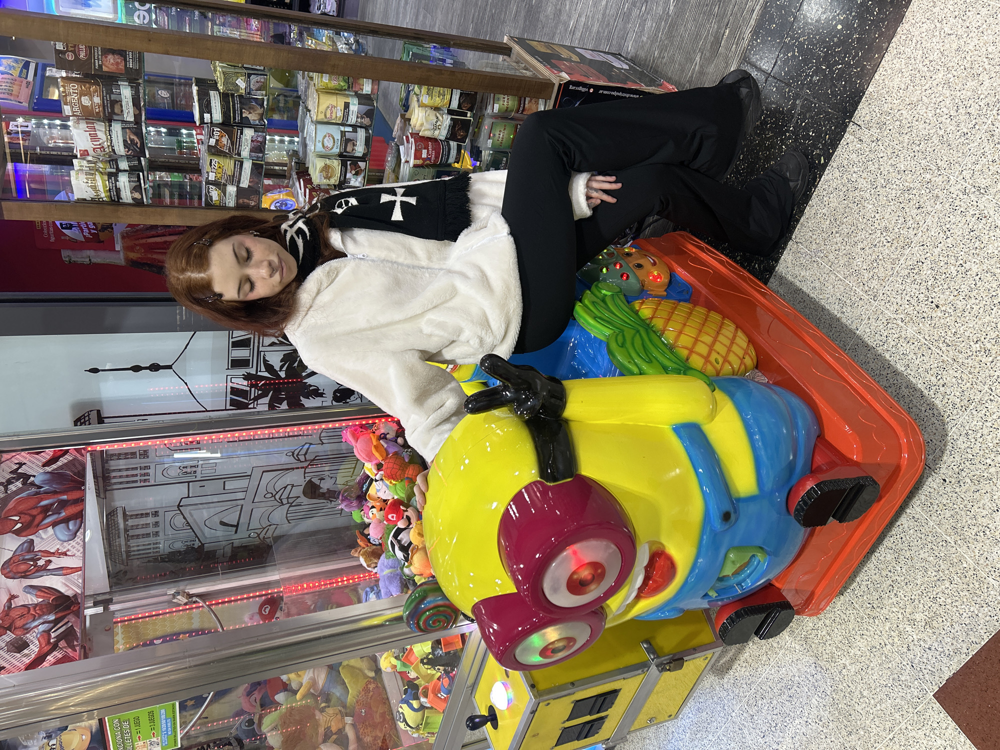 A young woman with auburn hair and wearing a white fluffy jacket and black pants is sitting on a Minion-themed children's ride. The ride is red, yellow, and blue and has oversized red goggles. The woman is leaning against the side of the ride. The scene appears to be inside a shopping mall, as evidenced by the tiled floor and the rows of merchandise, possibly cigarettes, displayed on shelves behind her. To the left is a claw machine filled with plush toys, including one of Spiderman. On the machine is a sign in Spanish that says "Funciona con billetes de." The photo was taken inside of a shopping mall in the city of Buenos Aires, Argentina.