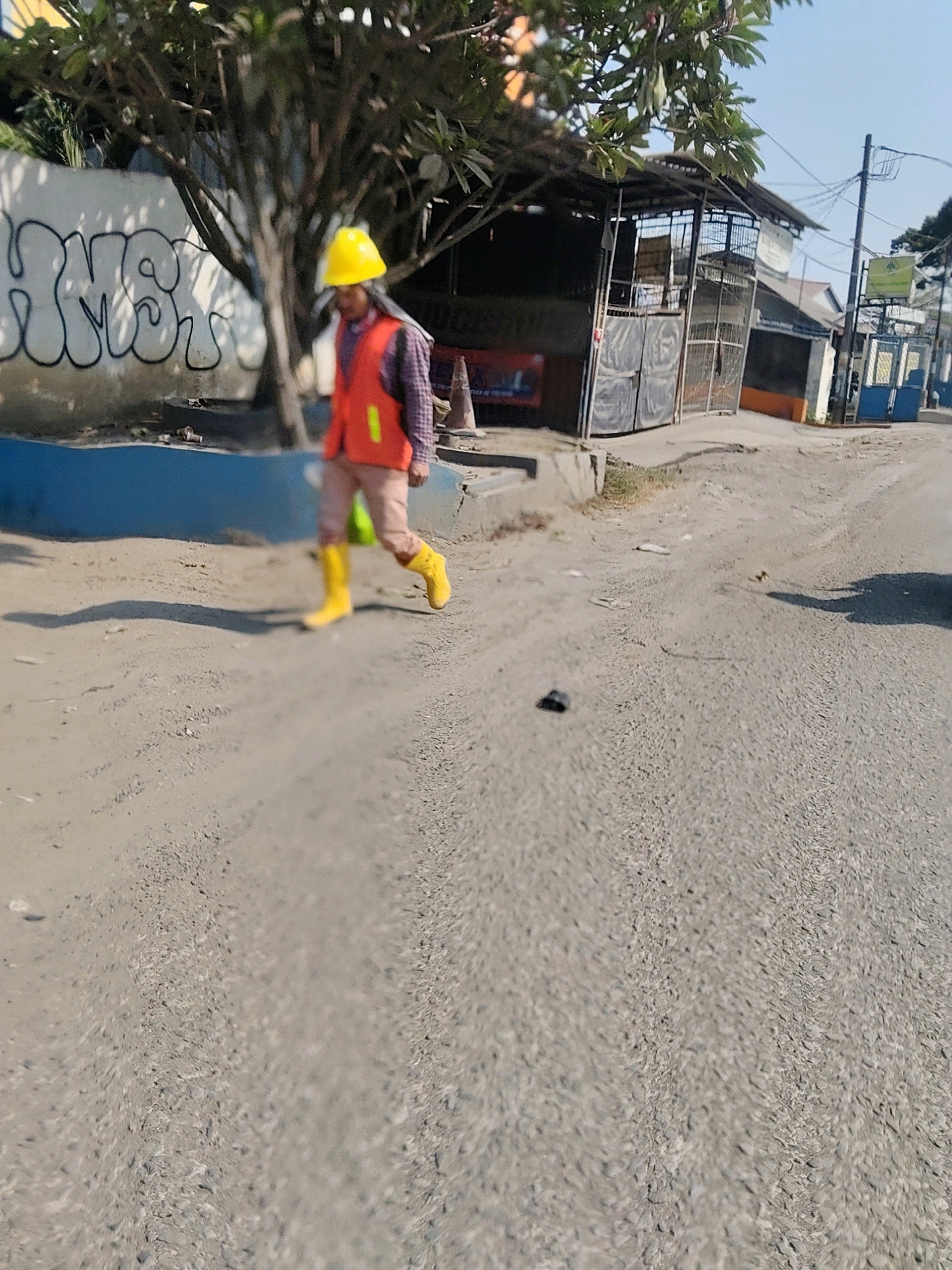 A person wearing a yellow hard hat and an orange reflective vest walks on a dusty, unpaved road. They are also wearing a plaid shirt and light-colored pants, along with bright yellow boots. The person appears to be moving from left to right in the frame. Behind them, a white wall with graffiti and a tree are visible. Further in the background, there is a building with a corrugated metal roof and open gates. The scene appears to be in an urban or semi-urban environment, characterized by the road, buildings, and utility poles. The lighting suggests it is daytime. The overall impression is one of a casual street scene, possibly in an area undergoing some form of construction or development. The specific location of Pasirsari, Indonesia, is indicated by the context provided.